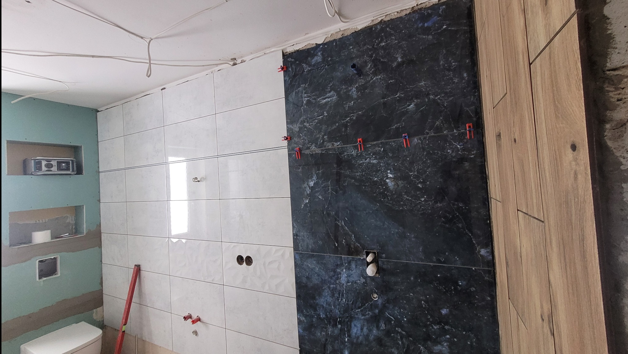 Wnętrze łazienki w trakcie układania płytek: białe, czarne marmurowe i drewnopodobne. Widoczne poziomica i klipsy do płytek. Częściowo wykończona ściana.