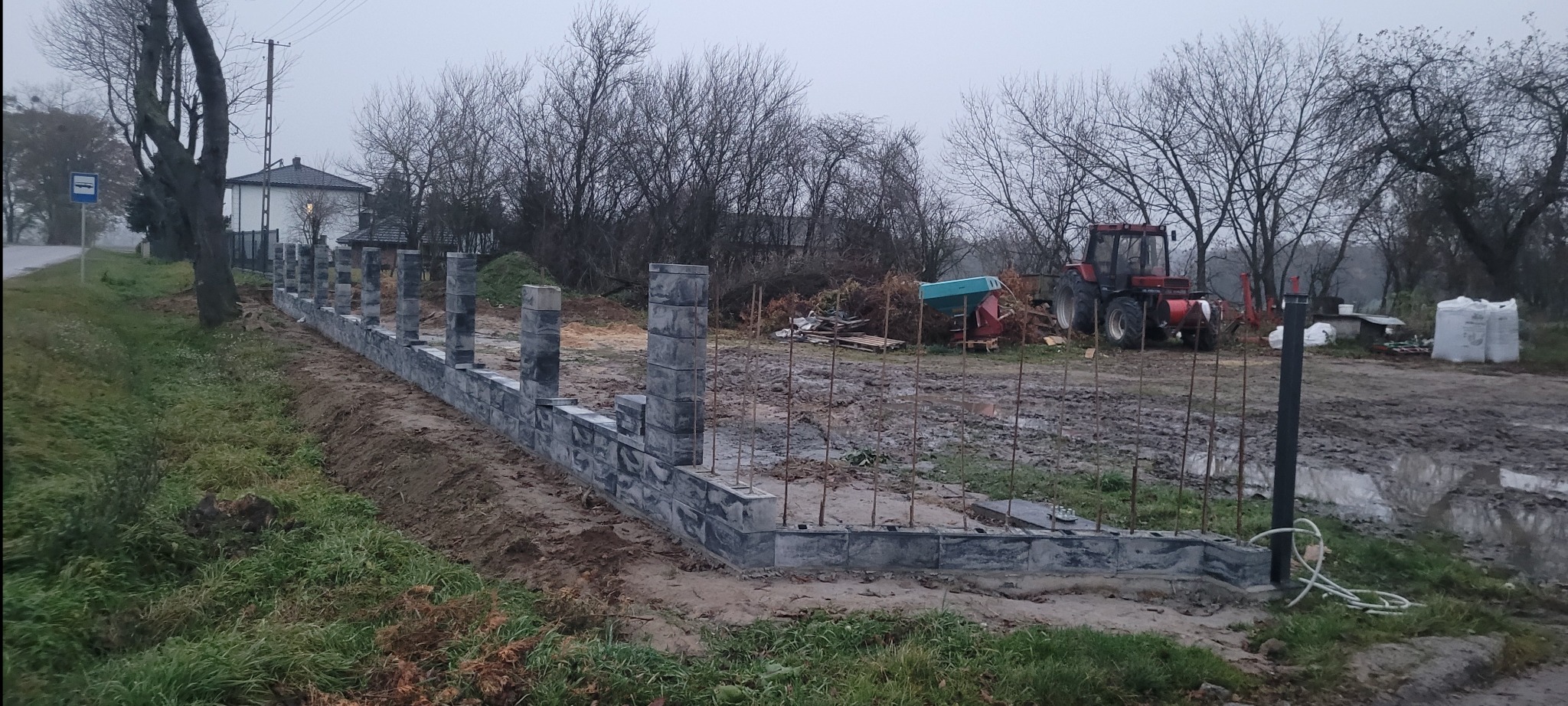Wznoszenie ogrodzenia z szarej kostki betonowej. Widoczne słupki i podmurówka w trakcie budowy. W tle traktor i teren budowy w pochmurny dzień.