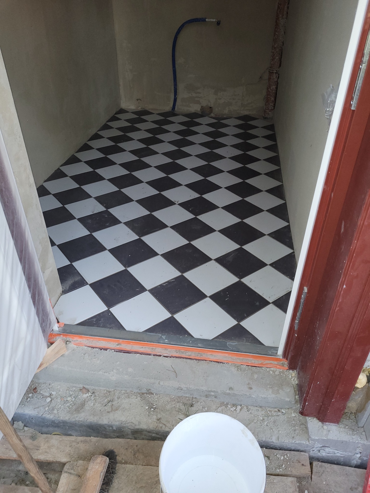 Ułożona podłoga z czarno-białych płytek w szachownicę w małym pomieszczeniu. Widoczny fragment drzwi i białe wiadro.