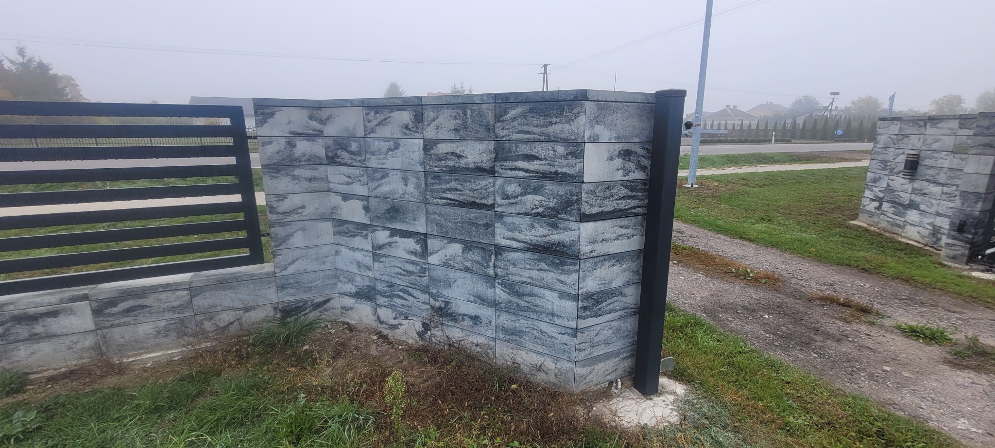 Nowoczesne ogrodzenie betonowe z elementami metalowymi w szarym odcieniu, widoczne z bliska w pochmurny dzień. Charakterystyczna faktura bloczków imitująca kamień.