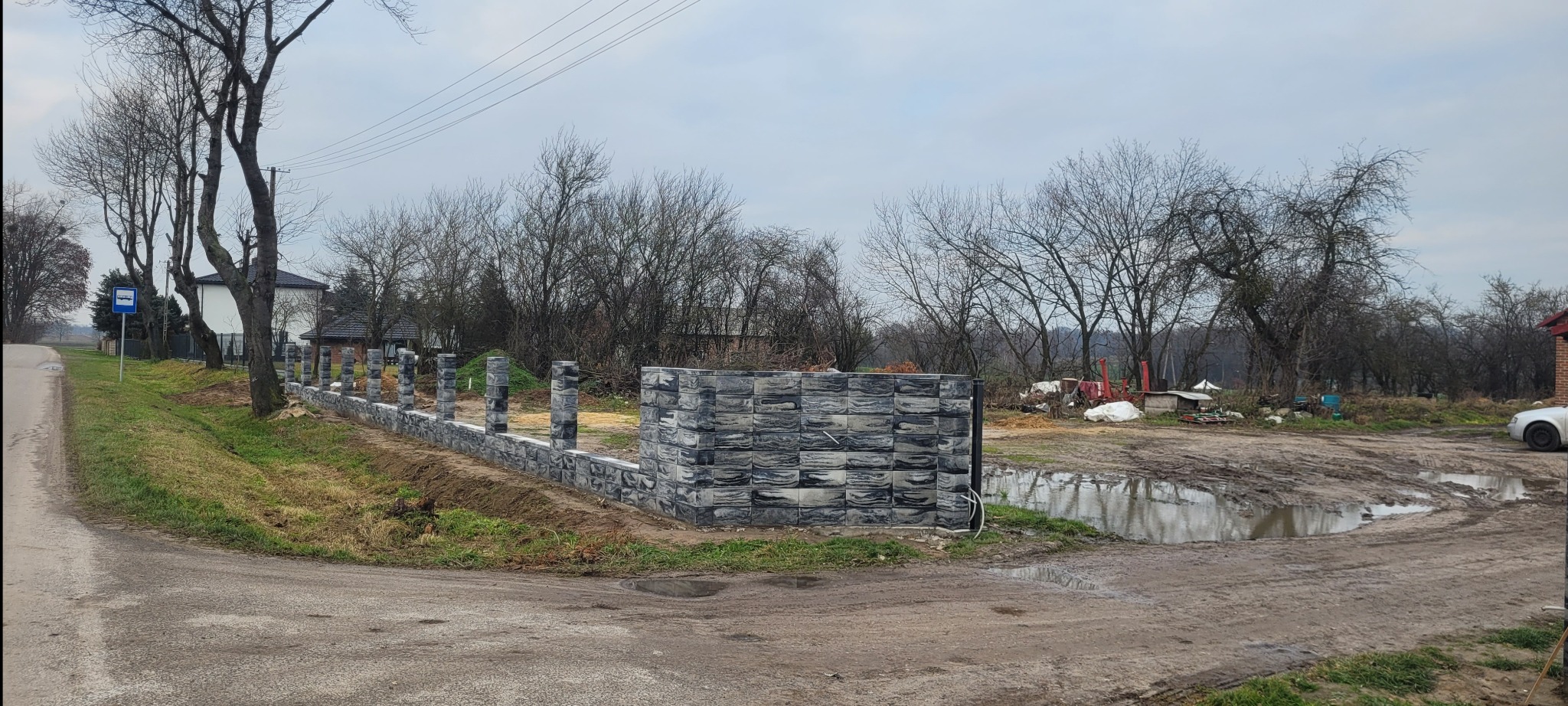 Fragment ogrodzenia betonowego z szarej kostki, widok z perspektywy ulicy w pochmurny dzień. Część ogrodzenia w trakcie budowy, widoczne słupki i fragment muru.
