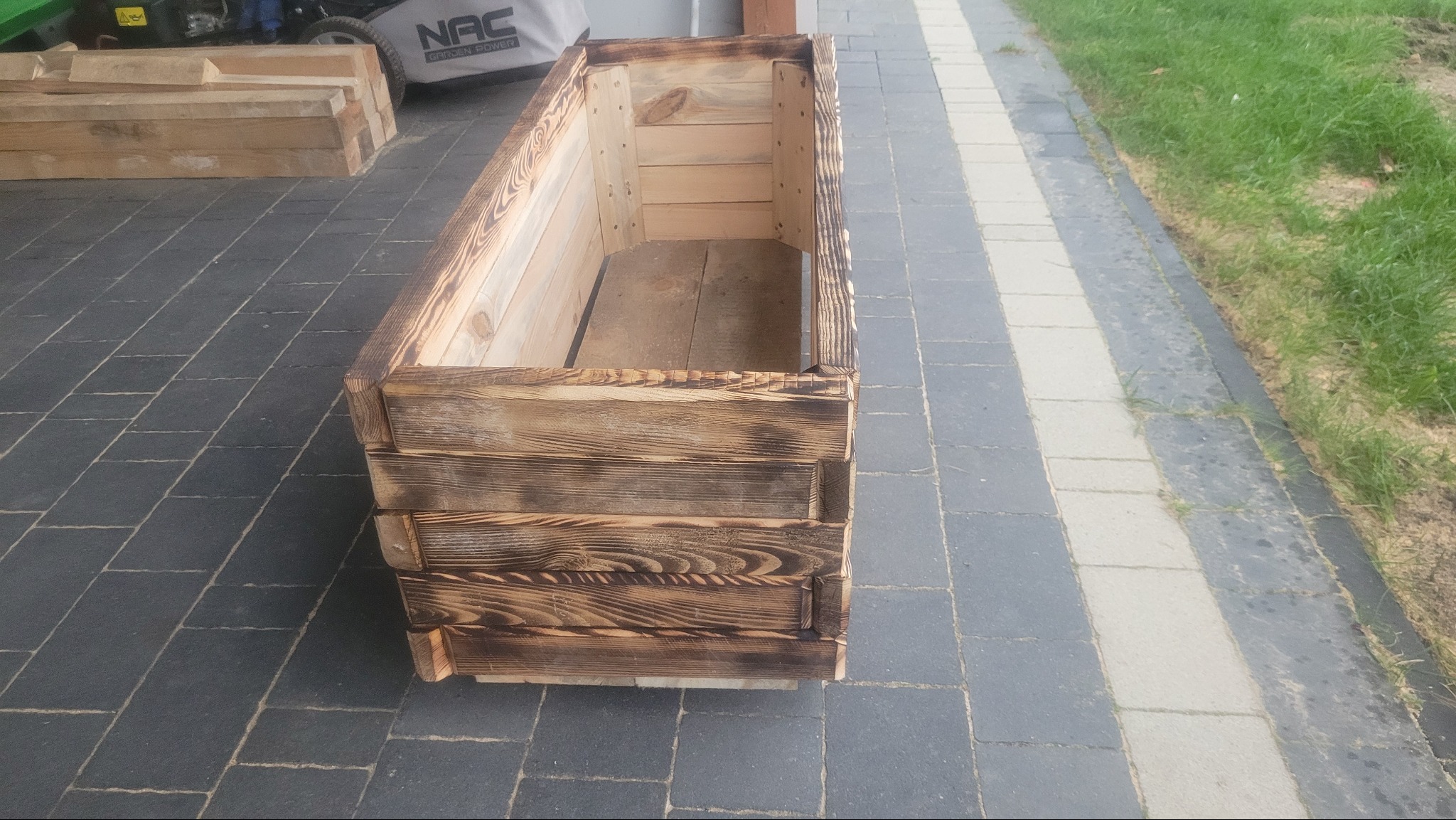 Drewniana skrzynia na kwiaty z opalonymi brzegami, ustawiona na szarym bruku. W tle trawnik i kosiarka. Widoczne detale konstrukcji i faktura drewna.