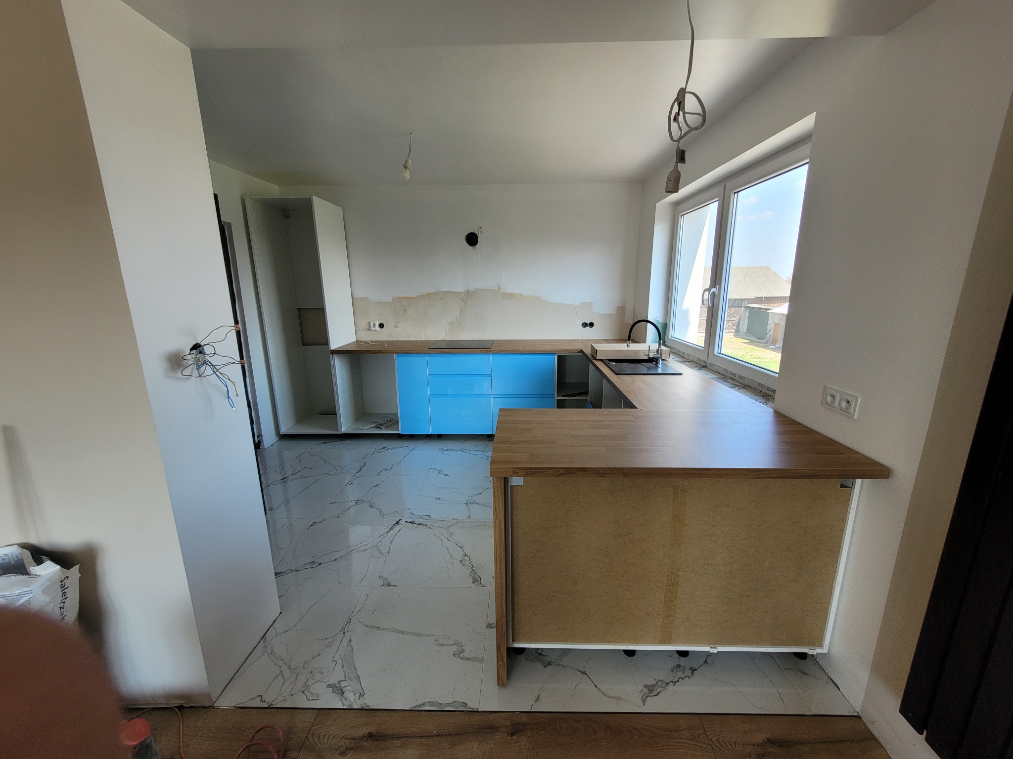 Wnętrze kuchni w trakcie montażu: niebieskie szafki, drewniany blat, marmurowe płytki podłogowe. Widoczne niedokończone ściany i instalacje elektryczne.