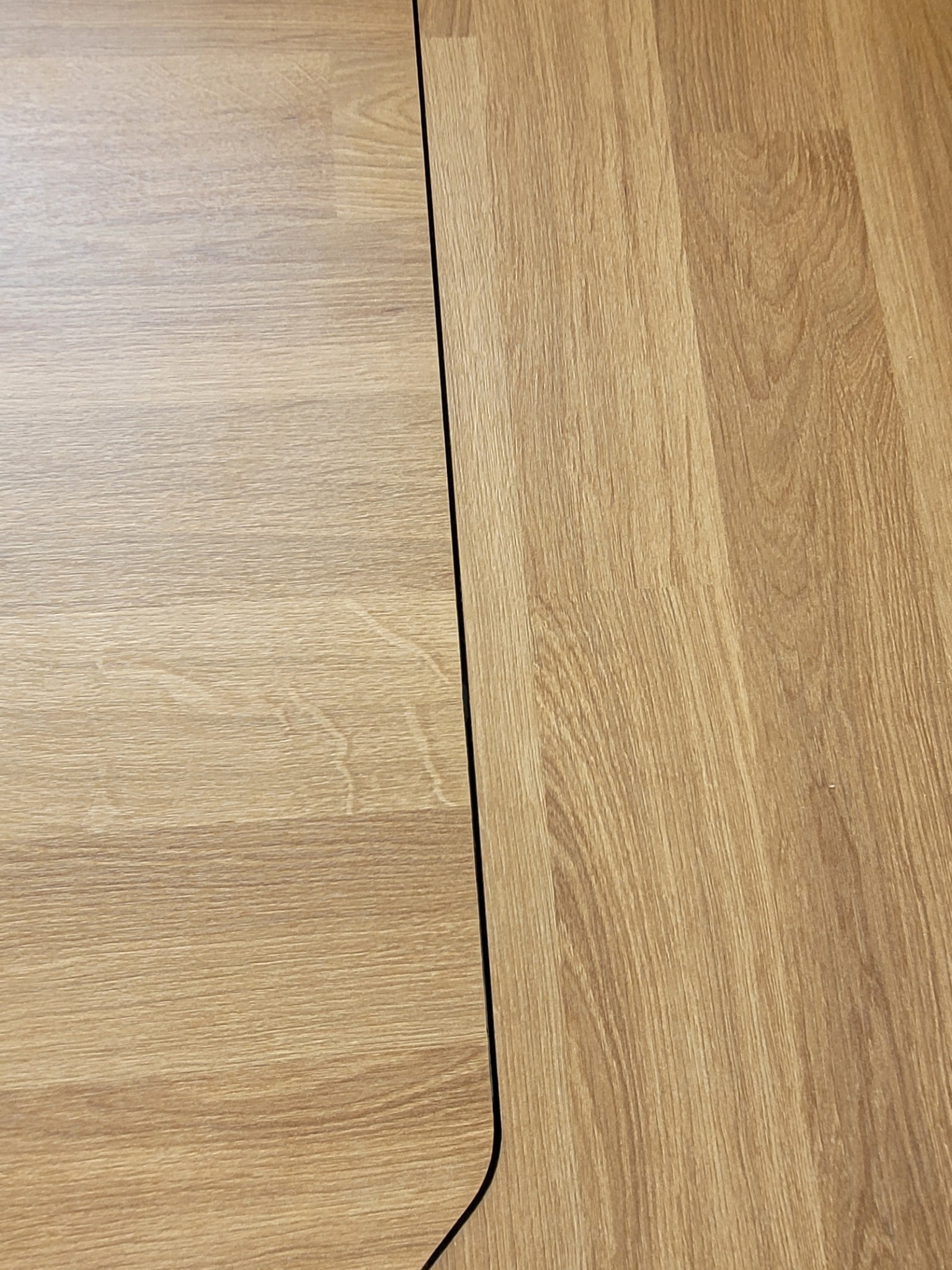 Jasne panele podłogowe z widoczną szczeliną dylatacyjną i zaokrąglonym narożnikiem. Układ pod kątem, imitacja drewna, ciepłe odcienie brązu i beżu.