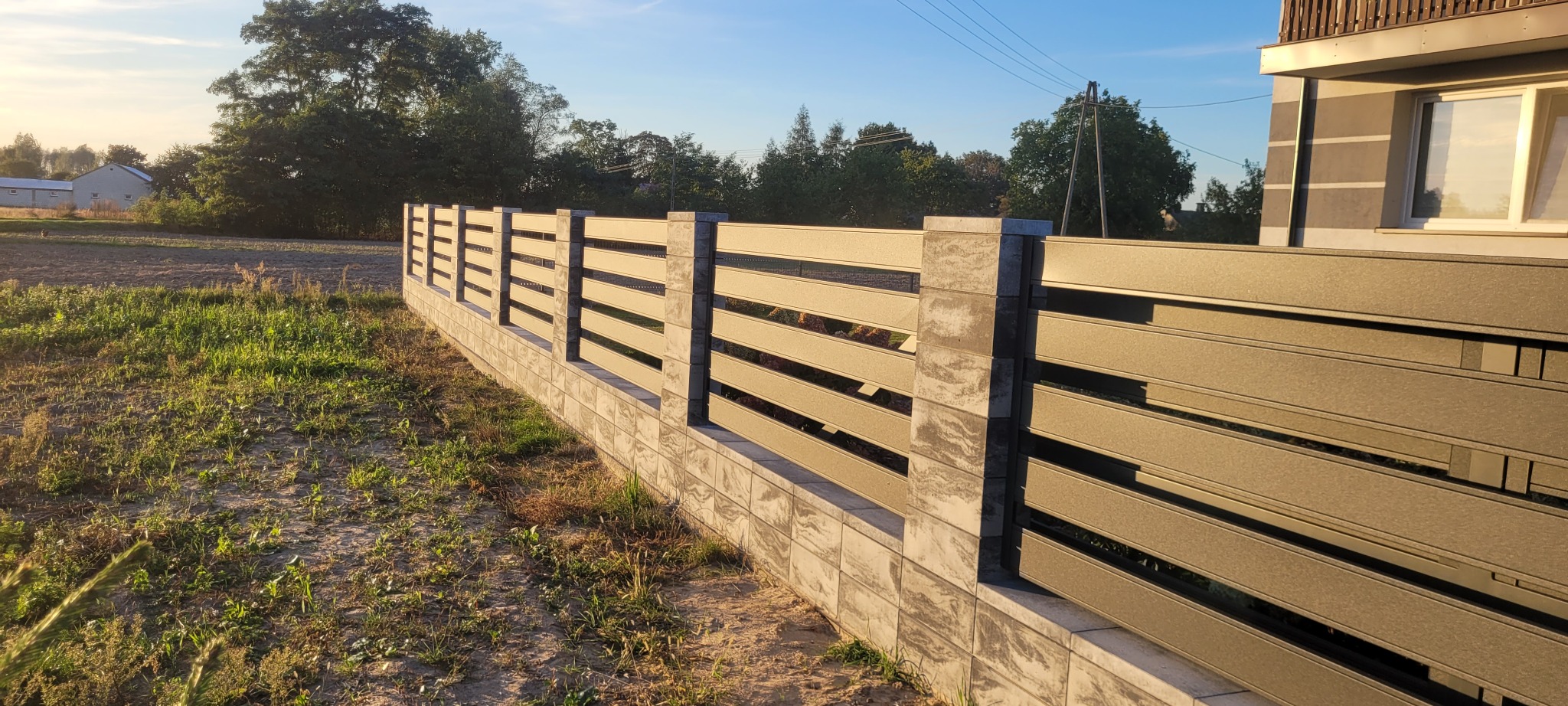 Nowoczesne ogrodzenie panelowe z betonową podmurówką, widok z boku na tle zieleni i budynku. Słoneczny dzień, wyraźne detale konstrukcji.