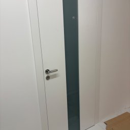 MERKANTOPRO SPÓŁKA Z OGRANICZONĄ ODPOWIEDZIALNOŚCIĄ - Białe drzwi wewnętrzne z matową szybą i srebrną klamką, widoczne na tle jasnej ściany i marmurowej podłogi. Minimalistyczny design.