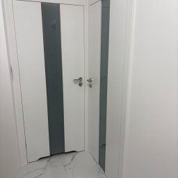 MERKANTOPRO SPÓŁKA Z OGRANICZONĄ ODPOWIEDZIALNOŚCIĄ - Dwa białe, nowoczesne drzwi z szarymi wstawkami, zamontowane w Dąbiu. Podłoga wyłożona białymi płytkami imitującymi marmur. Minimalistyczny design.