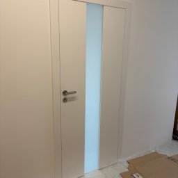 MERKANTOPRO SPÓŁKA Z OGRANICZONĄ ODPOWIEDZIALNOŚCIĄ - Białe, nowoczesne drzwi wewnętrzne z mleczną szybą pośrodku, zamontowane w jasnej ścianie. Na podłodze kartony zabezpieczające podczas prac wykończeniowych.