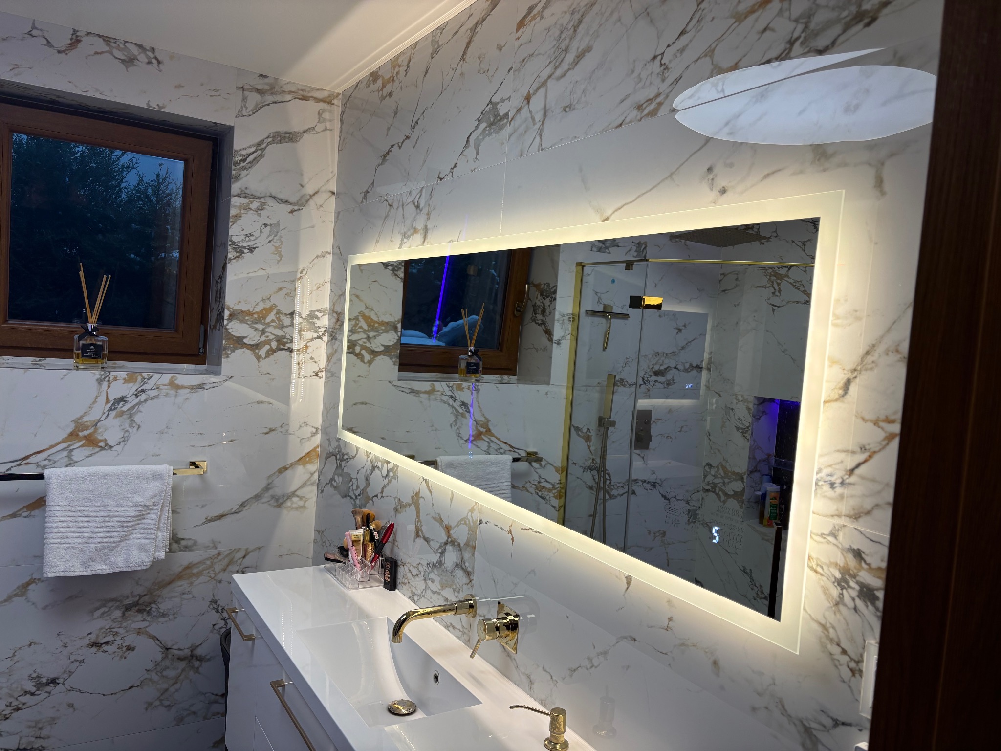 Elegancka łazienka z marmurowymi płytkami, podświetlanym lustrem i złotą armaturą. Widoczny fragment okna i akcesoria łazienkowe. Nowoczesny design.