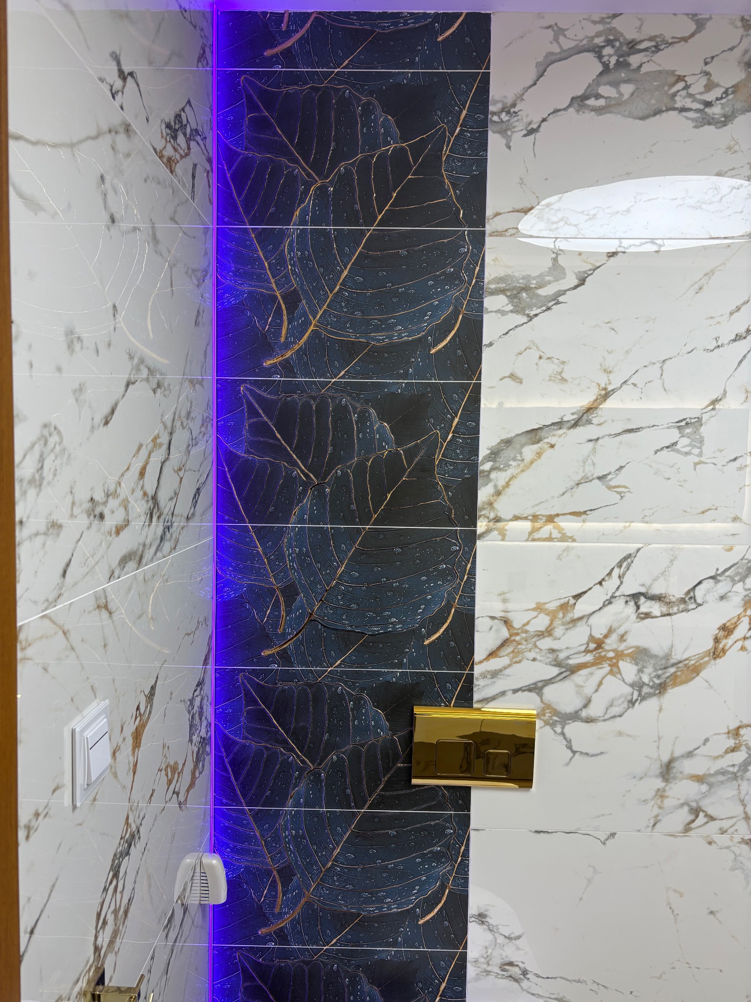 Wykończona ściana łazienki z płytek imitujących marmur i płytek z motywem liści, podświetlona niebieskim światłem LED. Widoczny złoty przycisk spłuczki.