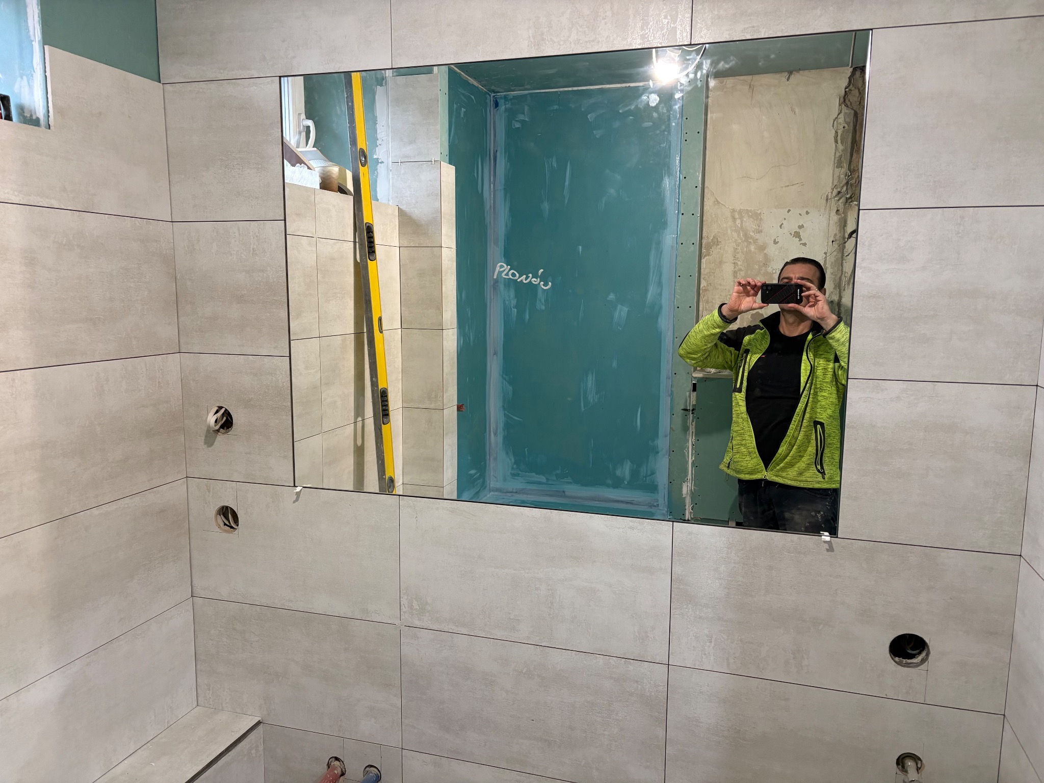 Łazienka w trakcie wykańczania: jasne, poziome płytki, otwory na armaturę. W lustrze odbija się osoba robiąca zdjęcie i fragment przygotowanej ściany pod dalsze prace.