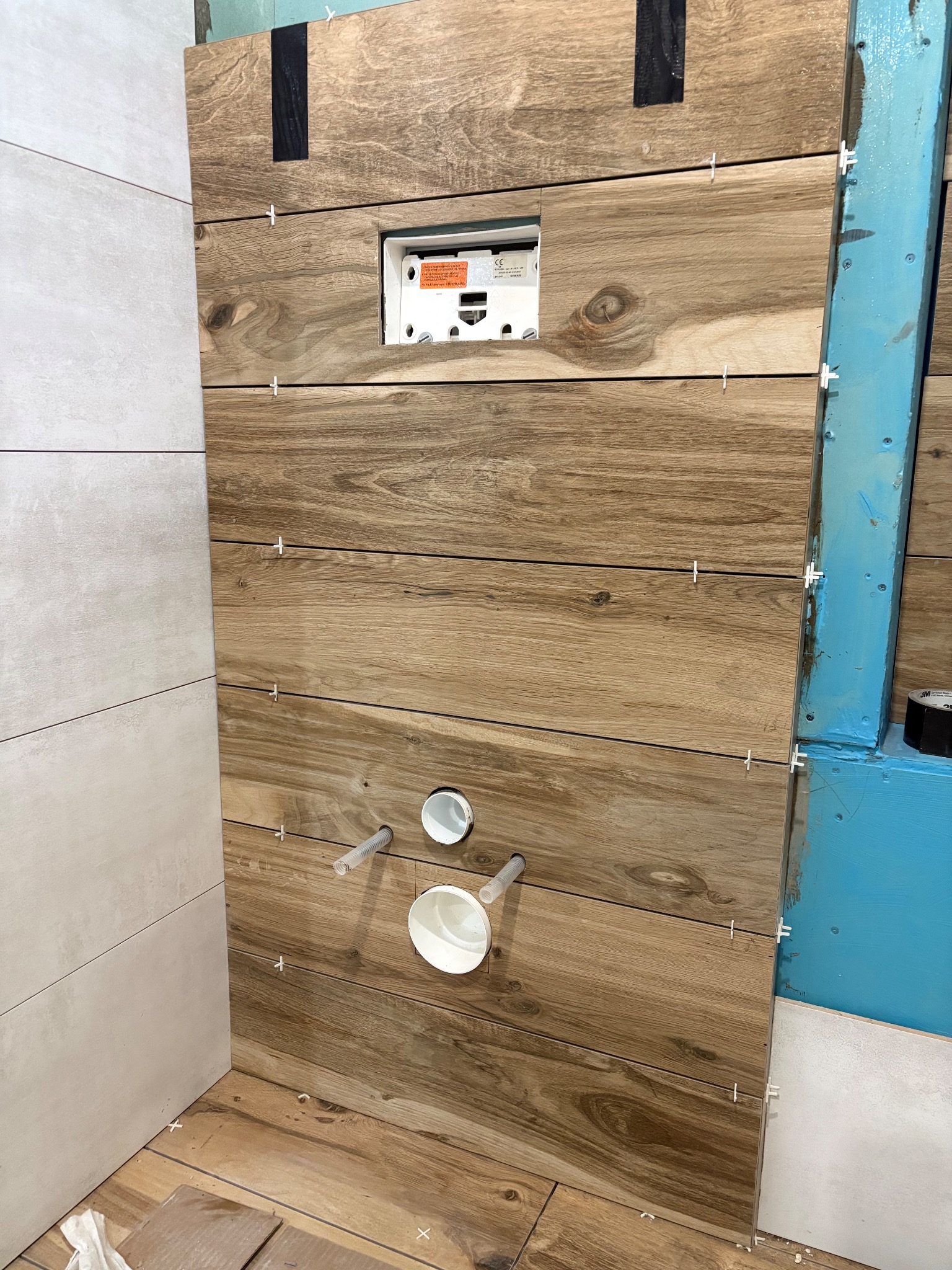 Ściana w łazience wyłożona płytkami imitującymi drewno, z widocznymi otworami na instalacje i krzyżykami dystansowymi, w trakcie prac wykończeniowych.