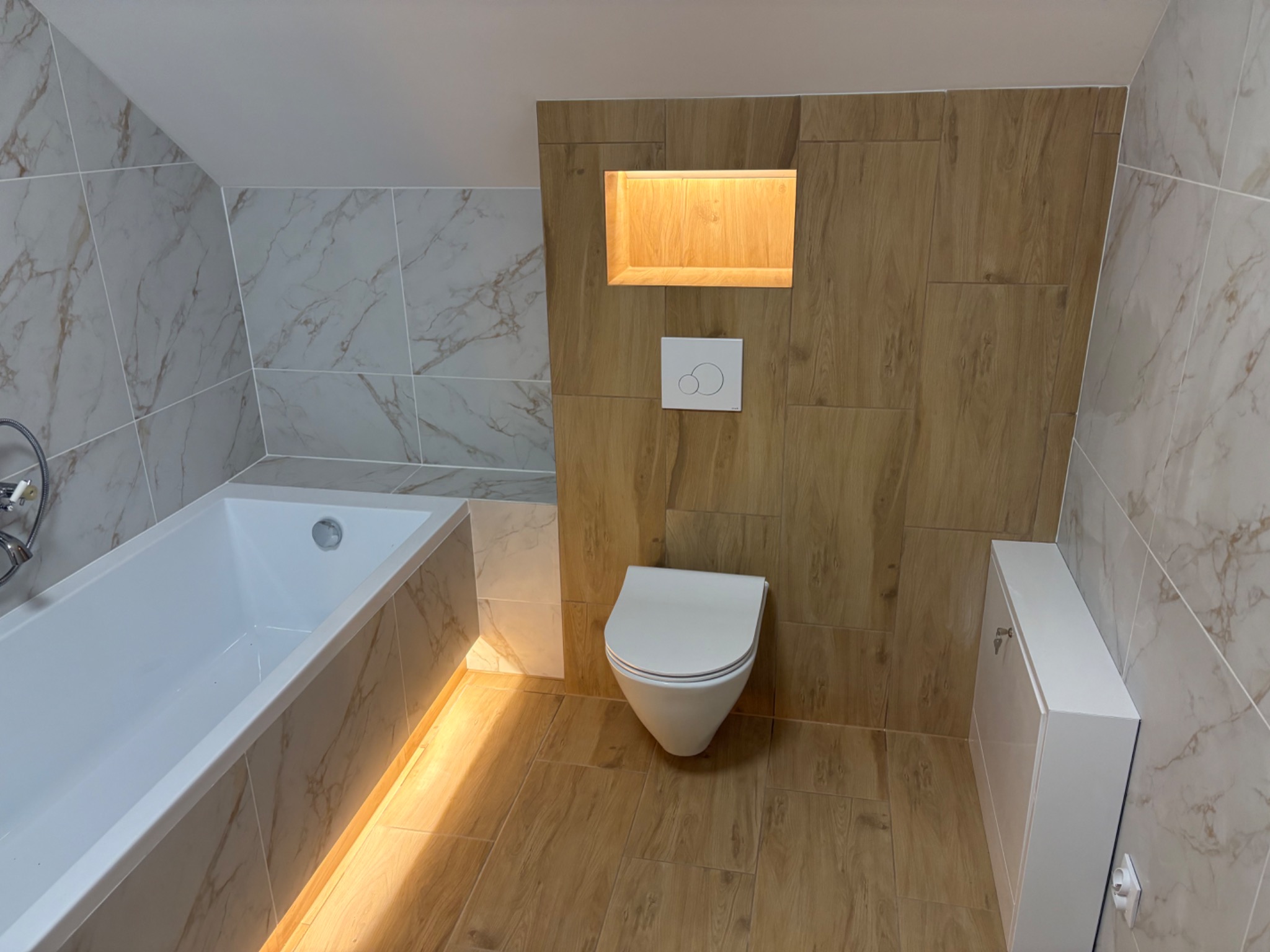 Nowoczesna łazienka na poddaszu z wanną, toaletą podwieszaną i drewnopodobnymi płytkami. Wnęka z oświetleniem nad toaletą. Marmurowe płytki na ścianach.