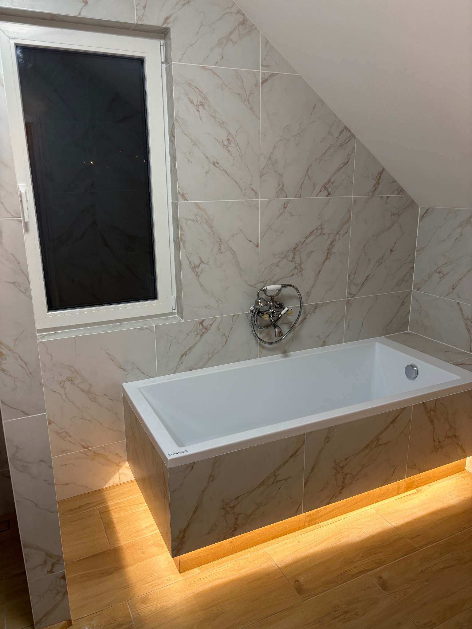 Biała wanna obudowana marmurowymi płytkami w łazience na poddaszu, z oknem i podświetleniem LED pod wanną, tworzącym ciepłą atmosferę. Bateria retro.