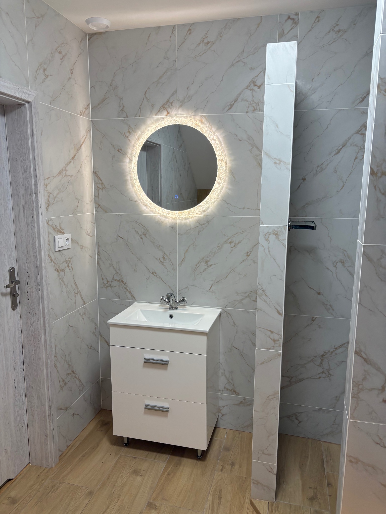 Wykończona łazienka z okrągłym lustrem LED, białą szafką pod umywalką i płytkami imitującymi marmur na ścianach. Widoczny fragment kabiny prysznicowej i drewnopodobna podłoga.