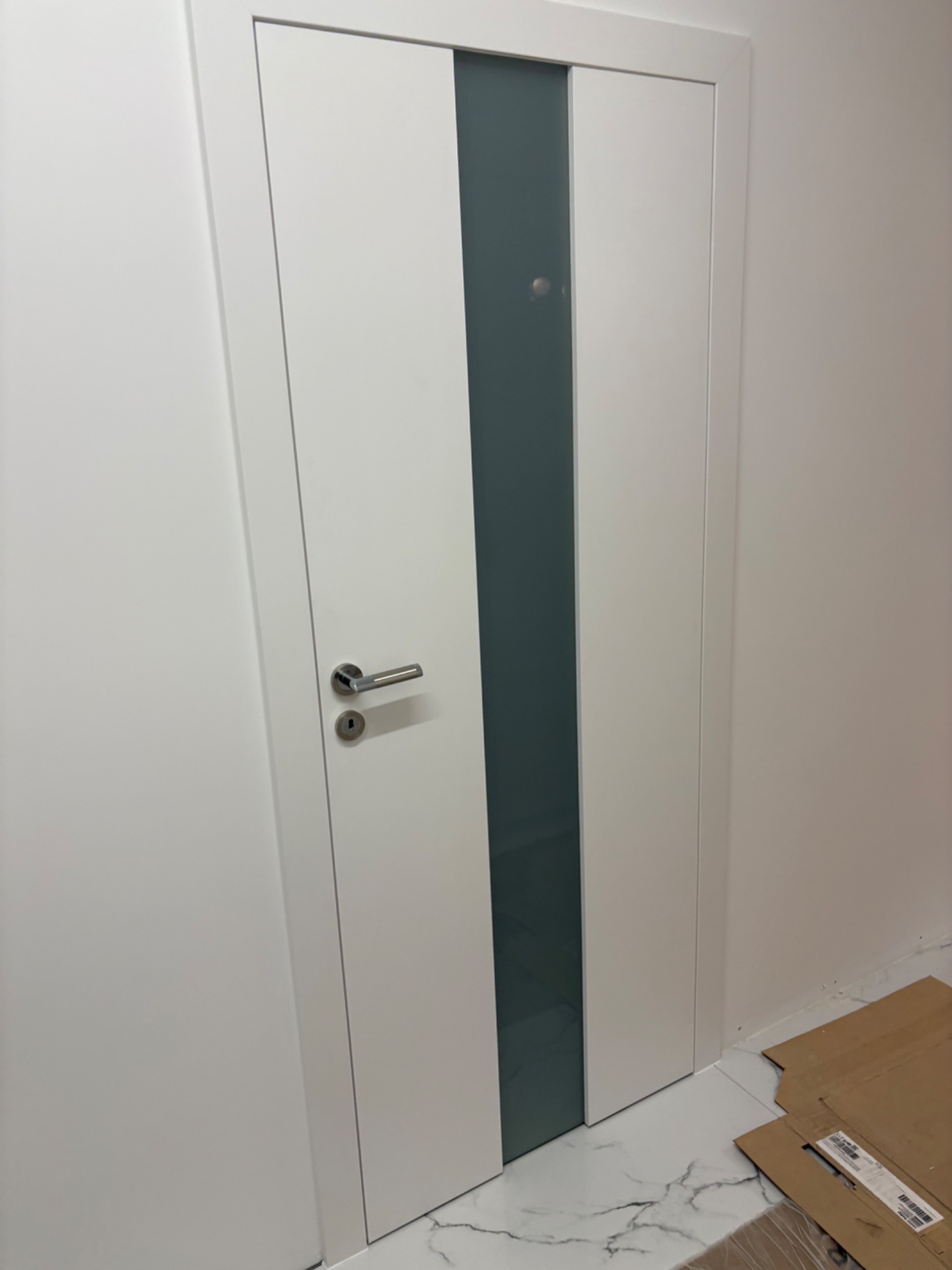 Białe drzwi wewnętrzne z matową szybą i srebrną klamką, widoczne na tle jasnej ściany i marmurowej podłogi. Minimalistyczny design.