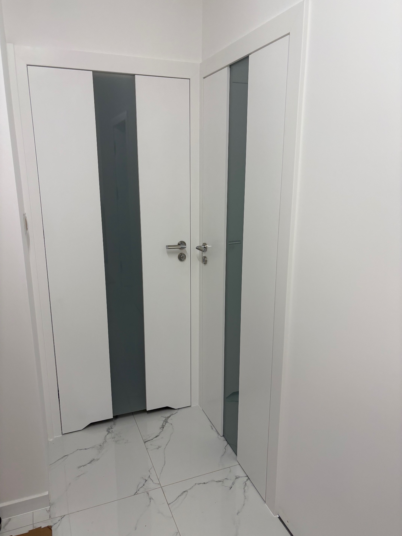 Dwa białe, nowoczesne drzwi z szarymi wstawkami, zamontowane w Dąbiu. Podłoga wyłożona białymi płytkami imitującymi marmur. Minimalistyczny design.