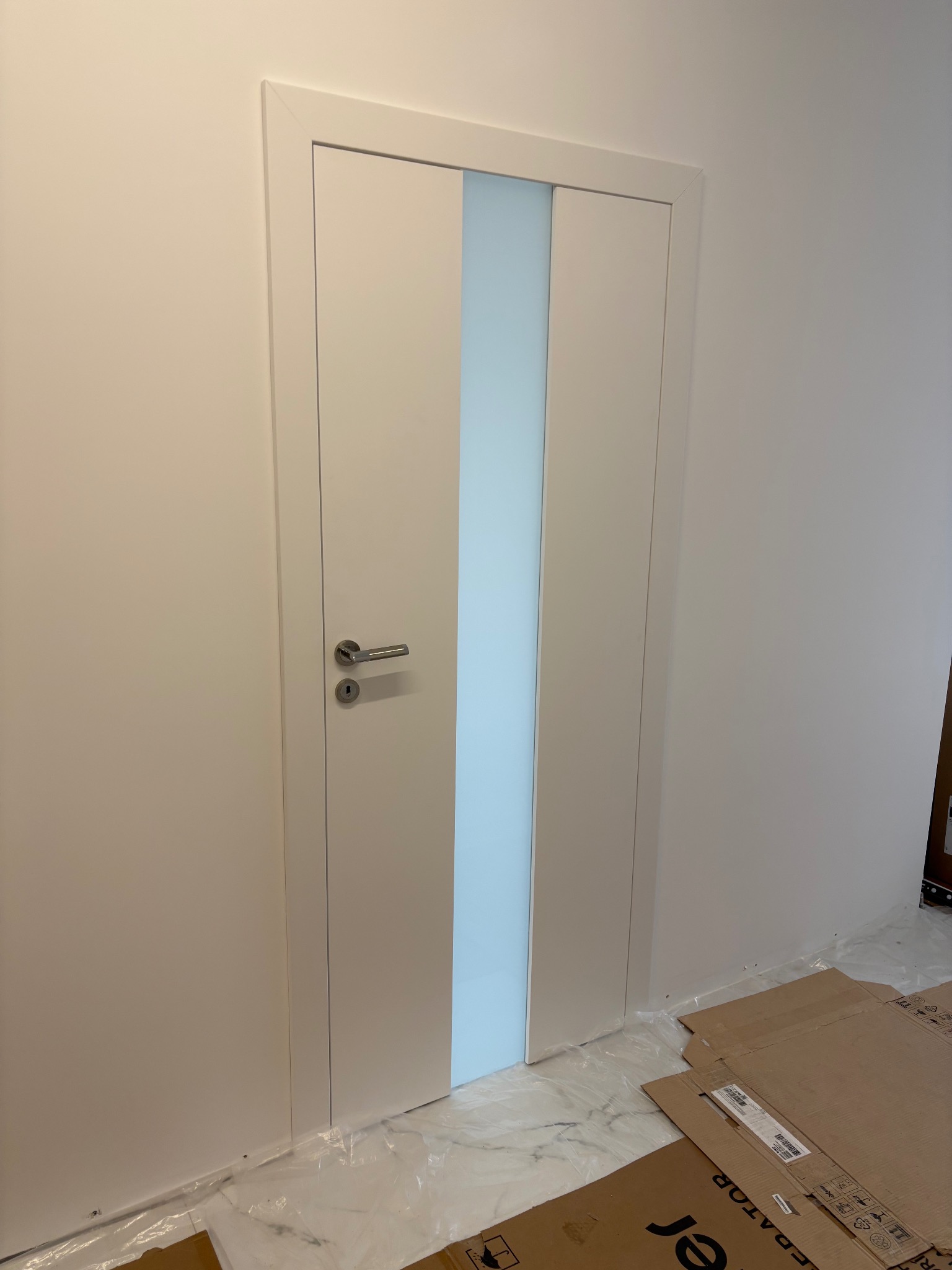 Białe, nowoczesne drzwi wewnętrzne z mleczną szybą pośrodku, zamontowane w jasnej ścianie. Na podłodze kartony zabezpieczające podczas prac wykończeniowych.