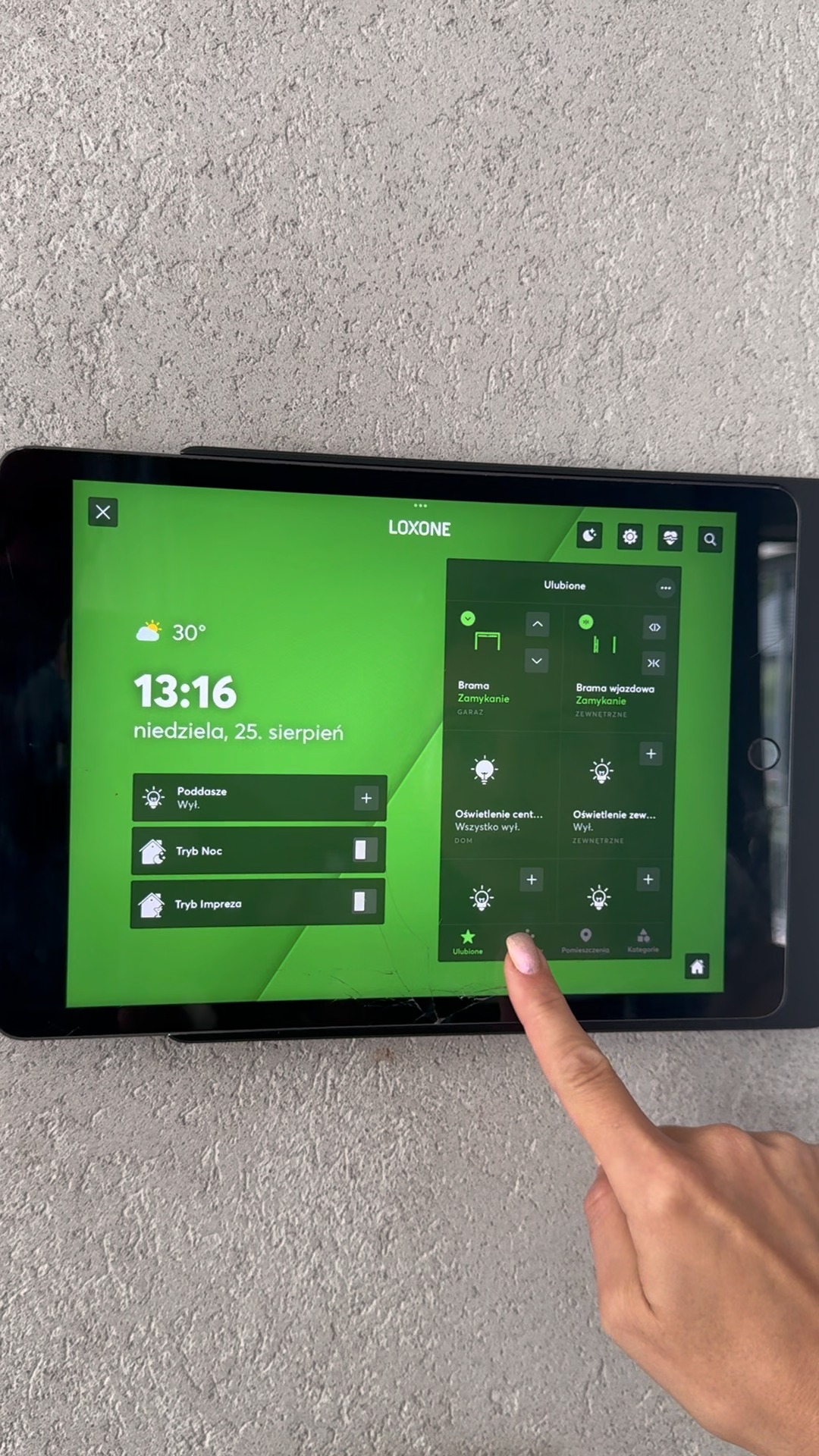 Tablet z aplikacją Loxone do sterowania inteligentnym domem, z widocznym interfejsem użytkownika i dłonią wskazującą na ekran. Widoczne opcje sterowania oświetleniem i bramą.