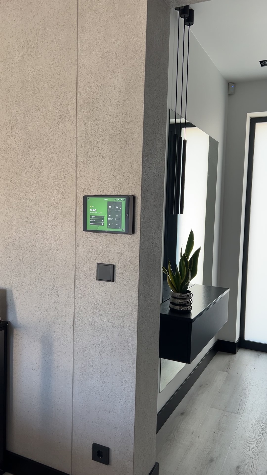 Panel sterowania inteligentnego domu zamontowany na ścianie obok włącznika i gniazdka. W tle lustro z konsolą i rośliną. Nowoczesne wykończenie wnętrza.