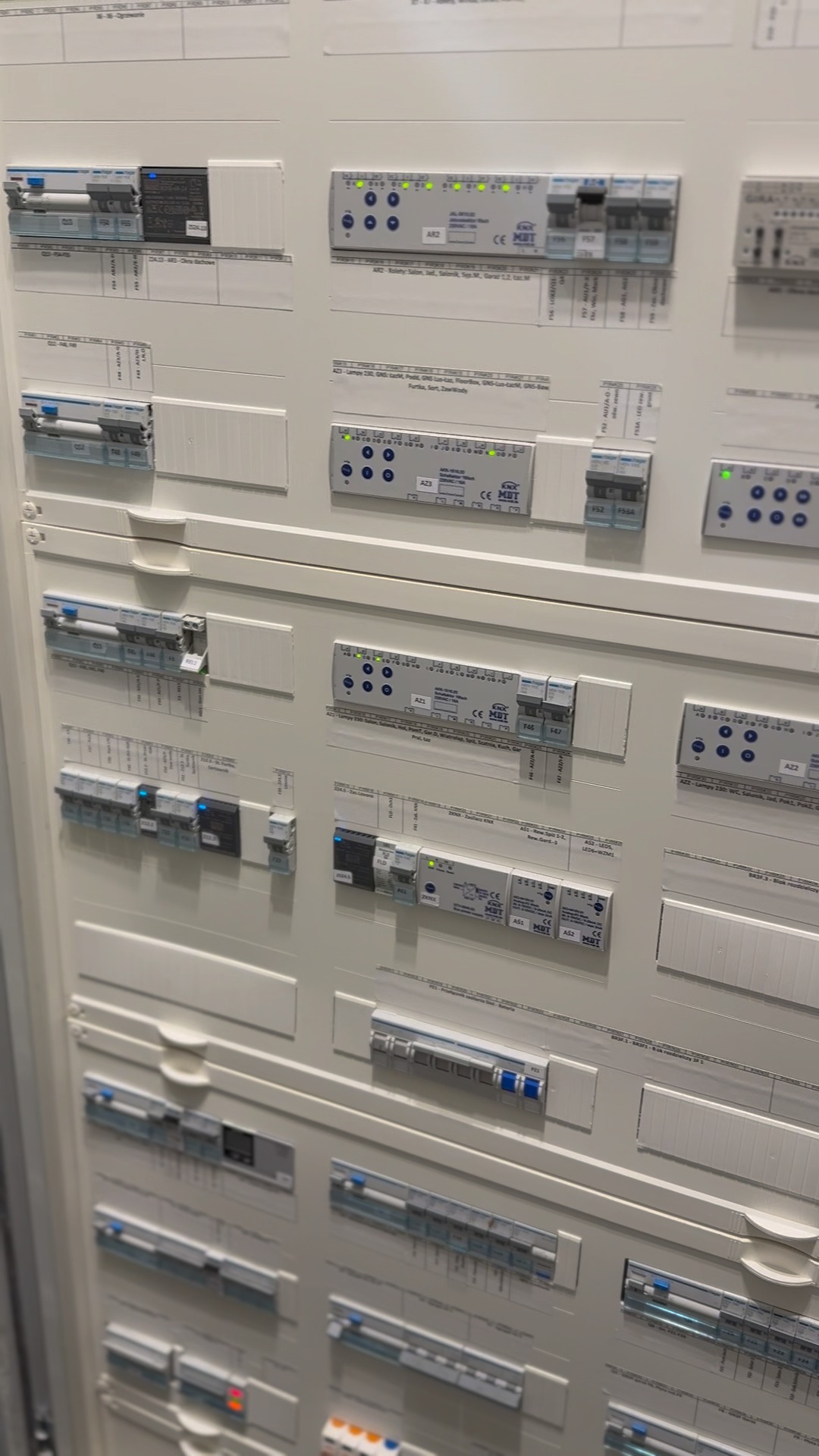 Rozdzielnica elektryczna z modułami sterującymi oświetleniem i roletami, widoczne oznaczenia i diody LED. Element instalacji inteligentnego domu.