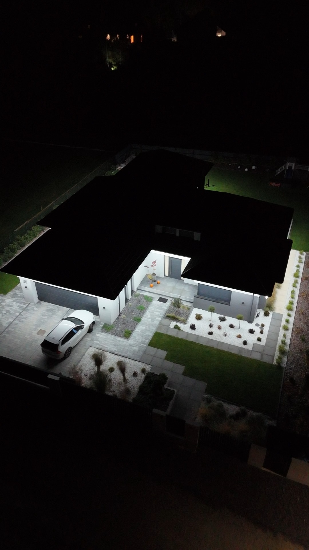 Nocny widok z góry na oświetlony dom z garażem i samochodem. Podświetlony ogród z białym żwirem i ścieżką z płyt. Nowoczesna architektura.