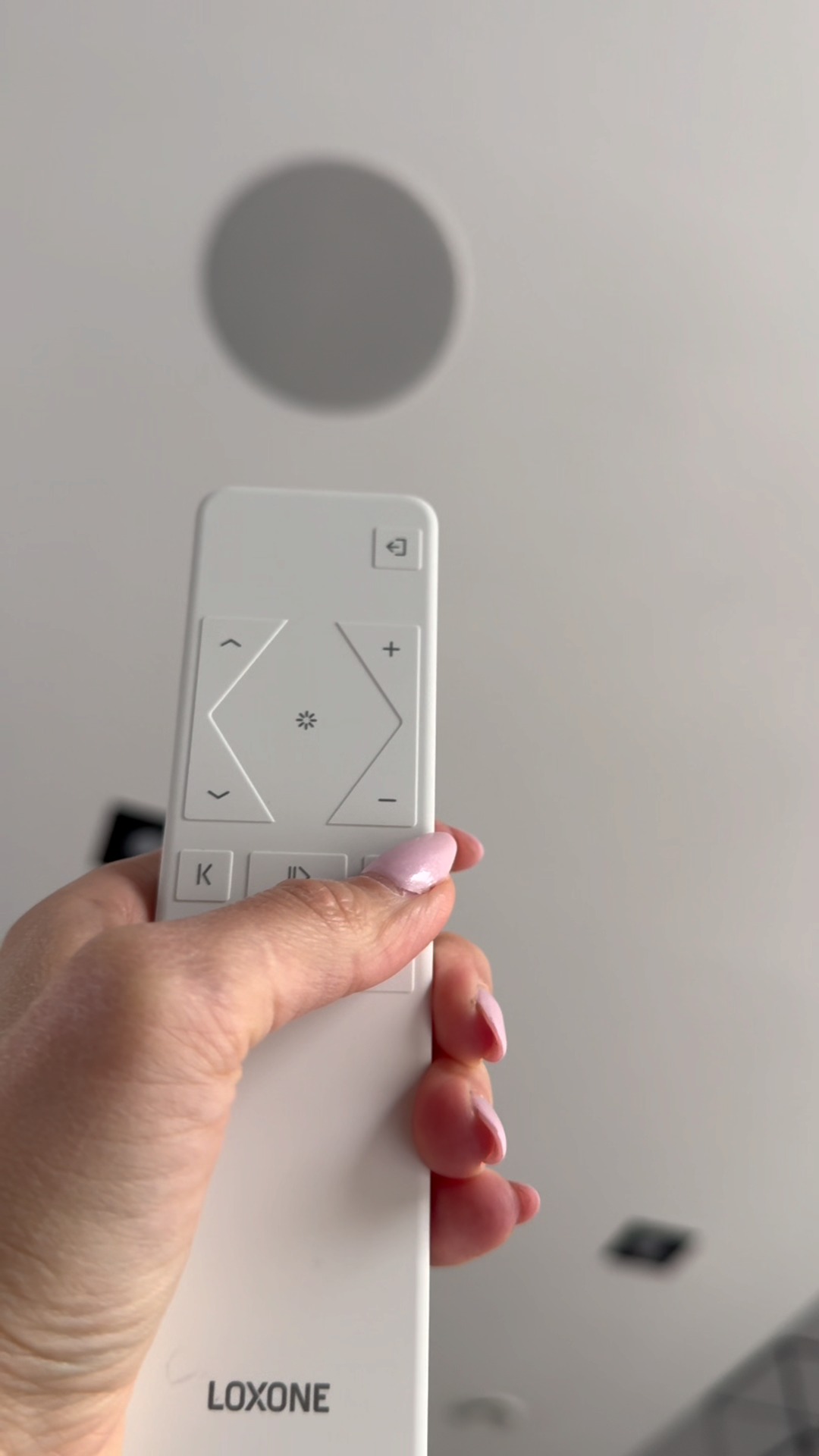 Biały pilot Loxone w dłoni kobiety, skierowany ku sufitowi z okrągłym głośnikiem. Sterowanie inteligentnym domem.
