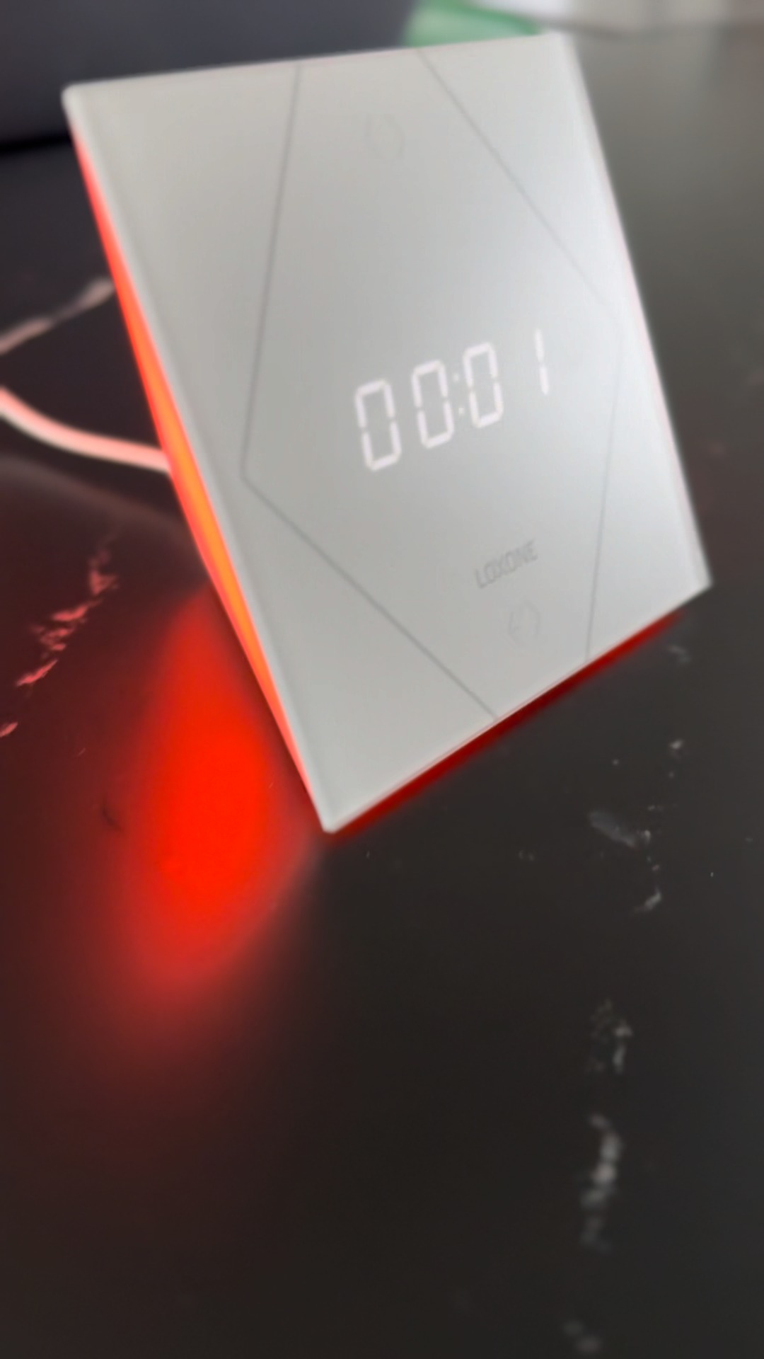 Biały panel sterowania Loxone z podświetleniem LED, wyświetlający godzinę 00:01, umieszczony na ciemnej, marmurkowej powierzchni. Widoczne delikatne odbicia światła.