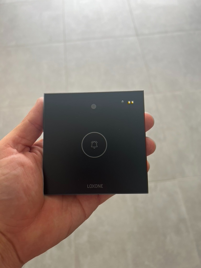 Czarny panel dotykowy Loxone z ikoną dzwonka, trzymany w dłoni na tle szarej podłogi. Widoczne diody LED i logo. Nowoczesny design, element inteligentnego domu.