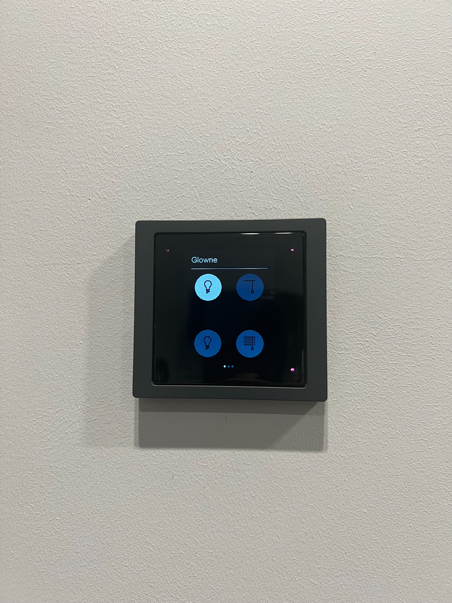 Panel sterowania inteligentnego domu, zamontowany na ścianie. Ekran dotykowy z ikonami: żarówka, lampa, roleta. Nowoczesny design, minimalistyczny styl.