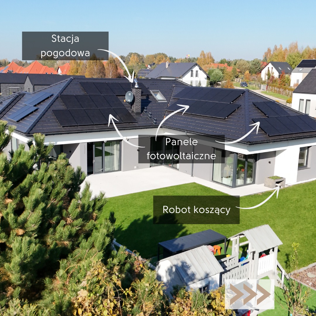 Nowoczesny dom jednorodzinny z panelami fotowoltaicznymi na dachu i stacją pogodową. Widoczny robot koszący na trawniku i plac zabaw. Ujęcie z góry, słoneczny dzień.