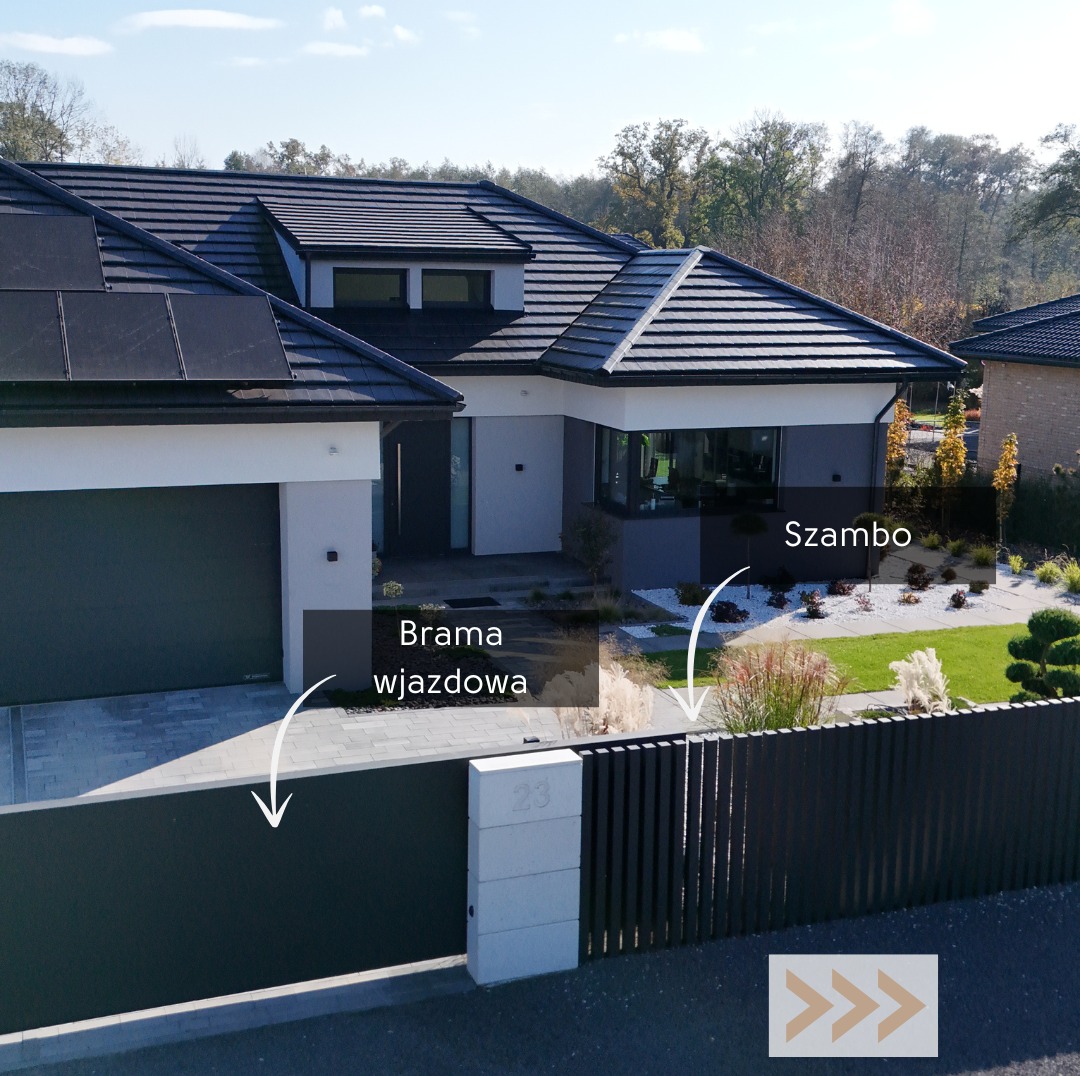 Nowoczesny dom jednorodzinny z szarą bramą wjazdową i panelami słonecznymi na dachu, otoczony zielenią i białymi kamieniami ozdobnymi.