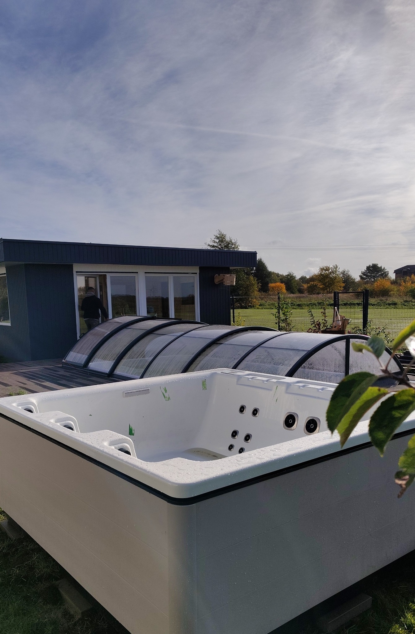 Nowoczesna wanna SPA na zewnątrz, zadaszona przeszkloną konstrukcją, obok budynku z dużymi oknami. Widok na ogród w tle, błękitne niebo z chmurami.