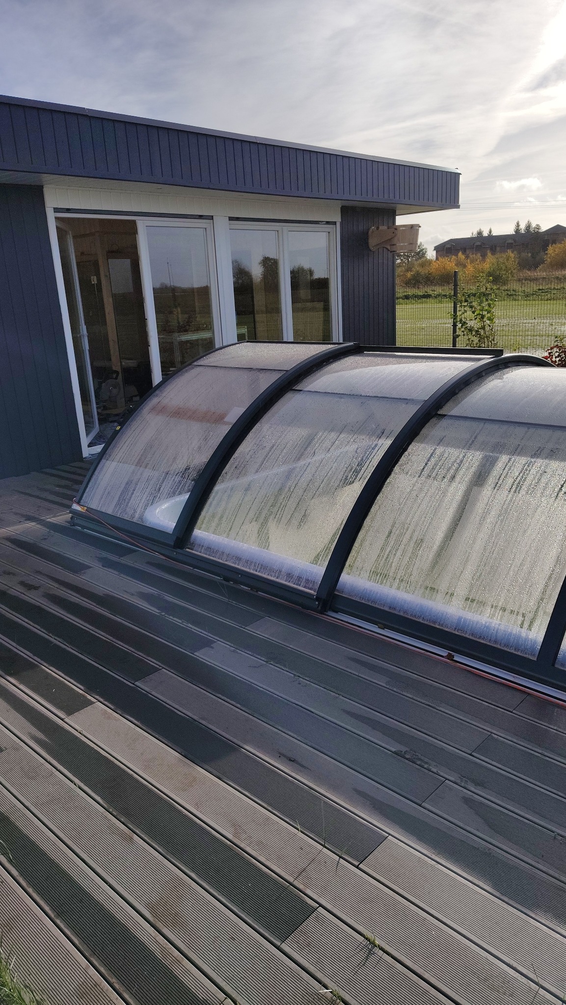 Zadaszenie basenu z poliwęglanu na drewnianym tarasie przy nowoczesnym domu. Widoczne krople wody na powierzchni. Tło stanowi zielony krajobraz.