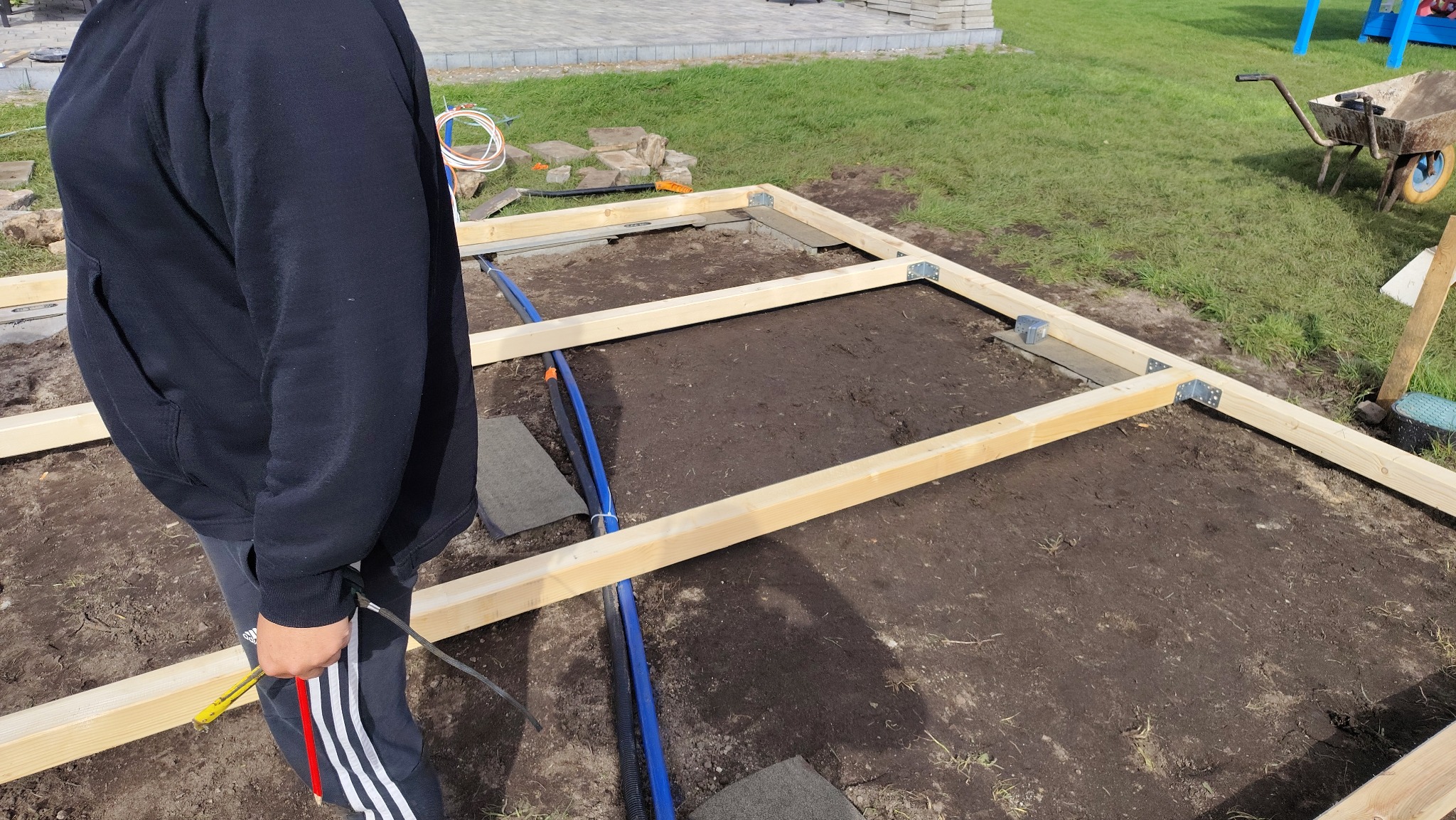 Konstrukcja z drewnianych belek na ziemi, prawdopodobnie pod taras lub altanę. Widoczne złącza kątowe, fragment osoby i narzędzia. Tło: trawa i elementy ogrodu.