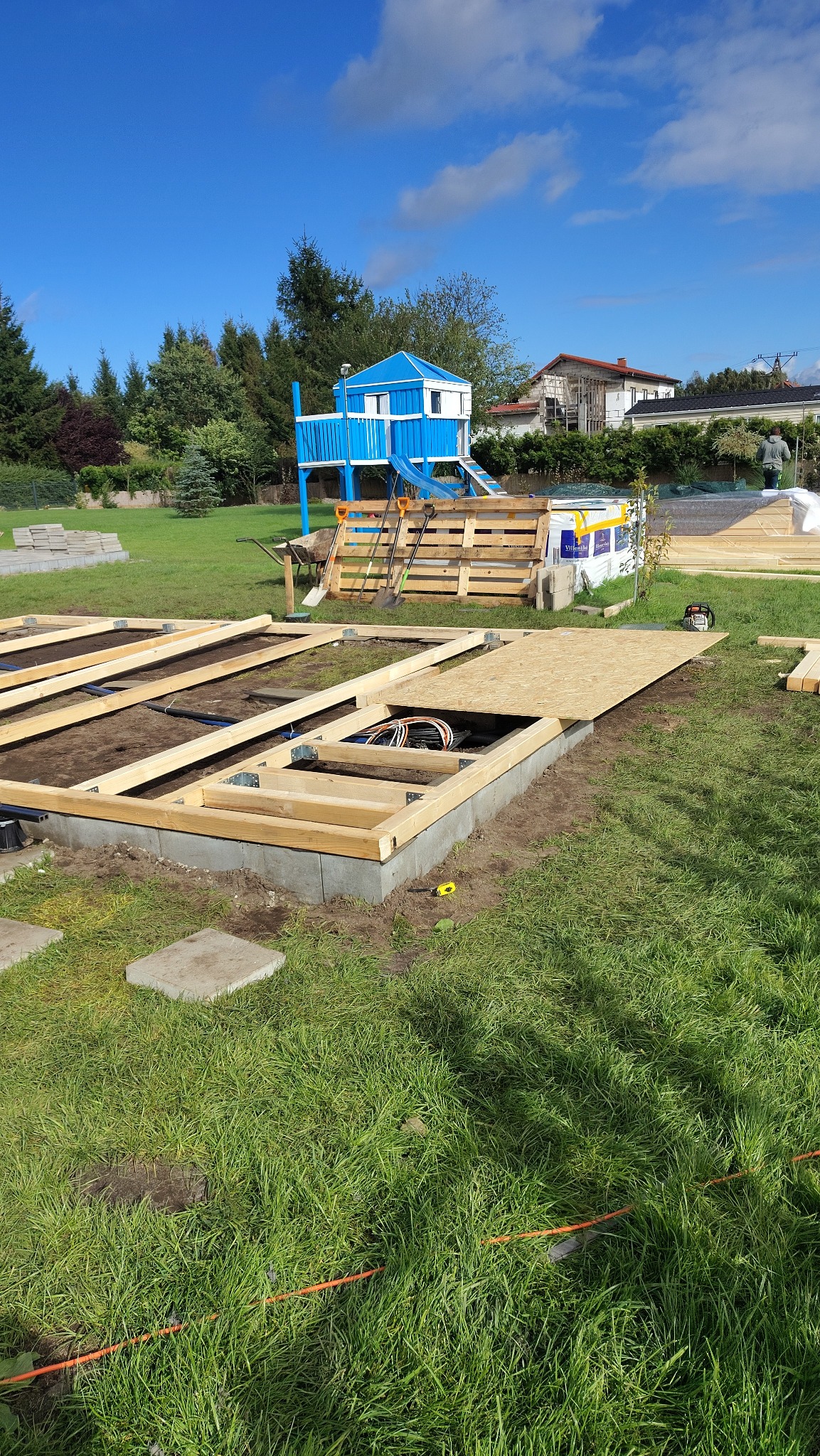 Budowa fundamentu pod domek ogrodowy z instalacją elektryczną. Widoczne drewniane ramy, płyta OSB i kable. W tle plac zabaw i zieleń.