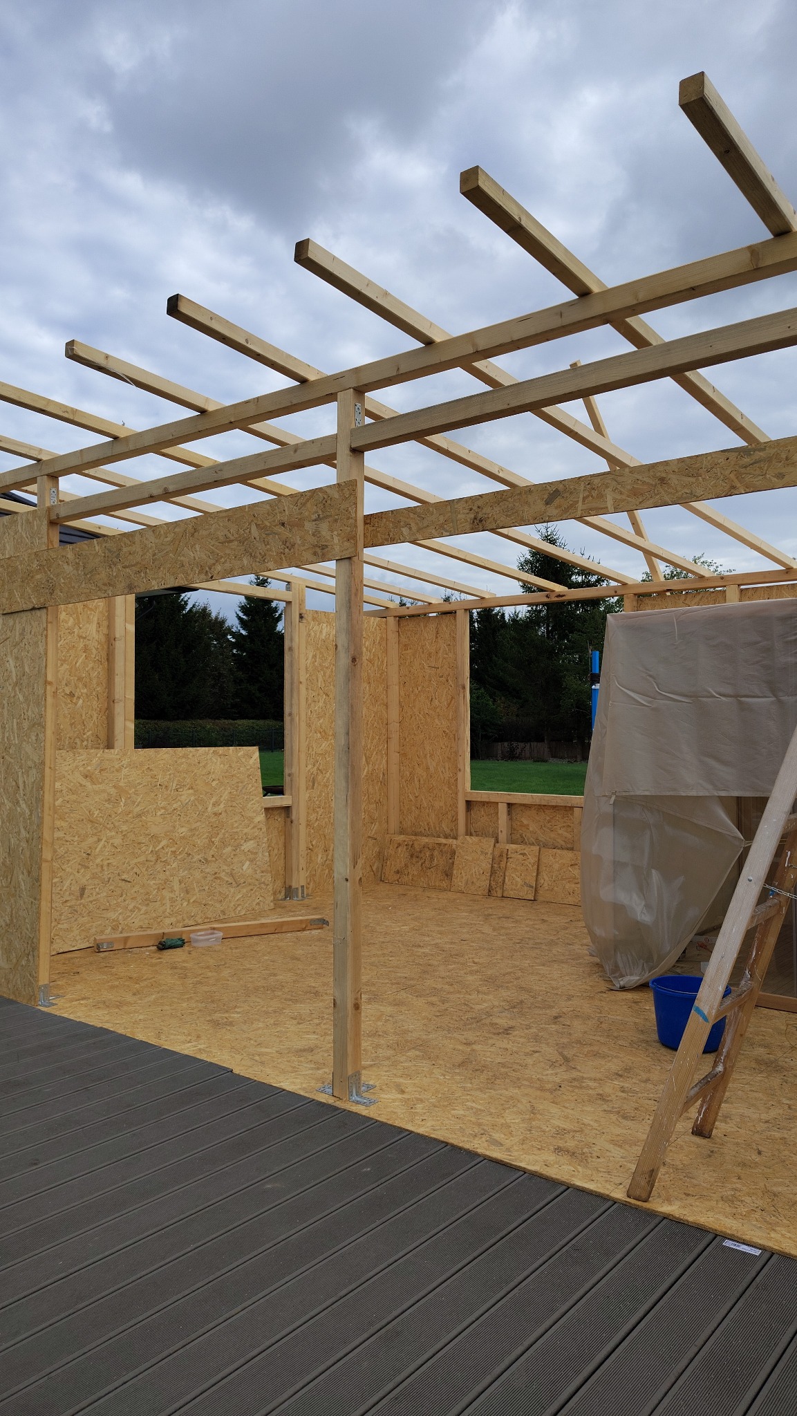 Konstrukcja altany ogrodowej w trakcie budowy: drewniany szkielet z płyt OSB, widoczne elementy mocujące, drabina i fragment tarasu kompozytowego. Pochmurne niebo w tle.