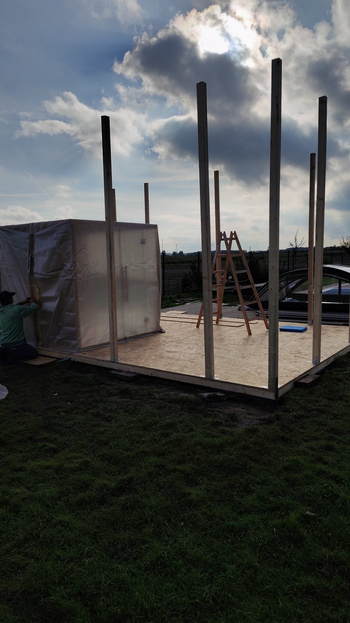 Budowa szkieletu drewnianego na płycie OSB, widoczne słupy i częściowo obudowana konstrukcja. W tle drabina i pochmurne niebo.