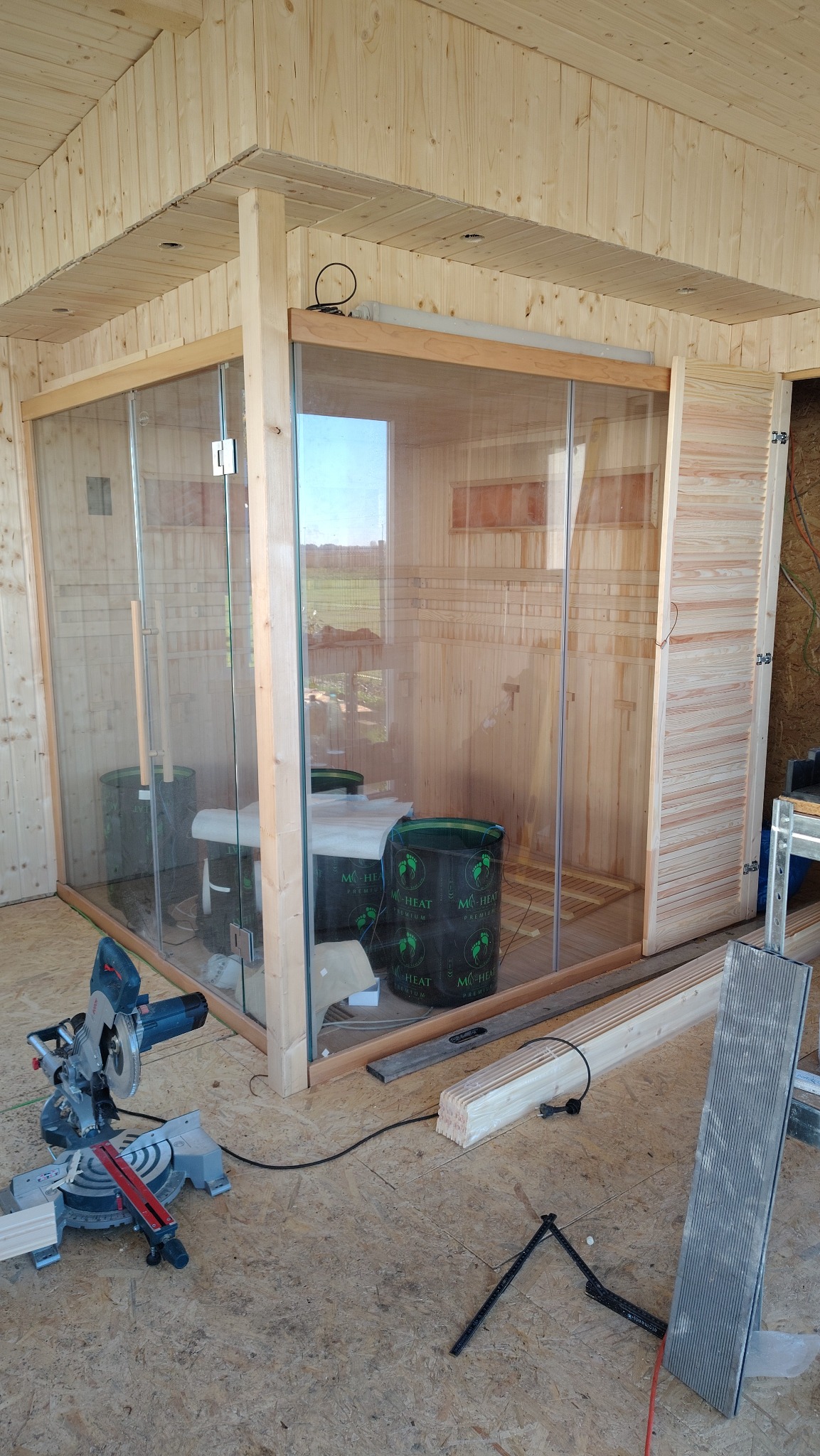 Wnętrze sauny w trakcie budowy: drewniane ściany, szklane drzwi, widoczne narzędzia (piła elektryczna) i materiały budowlane. W tle widok na zewnątrz.