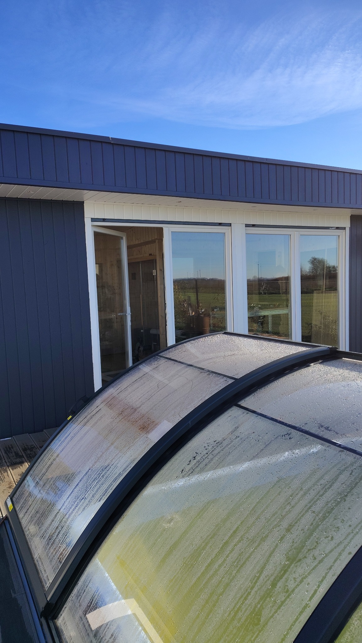 Szklana konstrukcja z kroplami wody, prawdopodobnie zadaszenie basenu, na tle budynku z dużymi oknami i błękitnym niebem. Widok z góry, jasne światło.