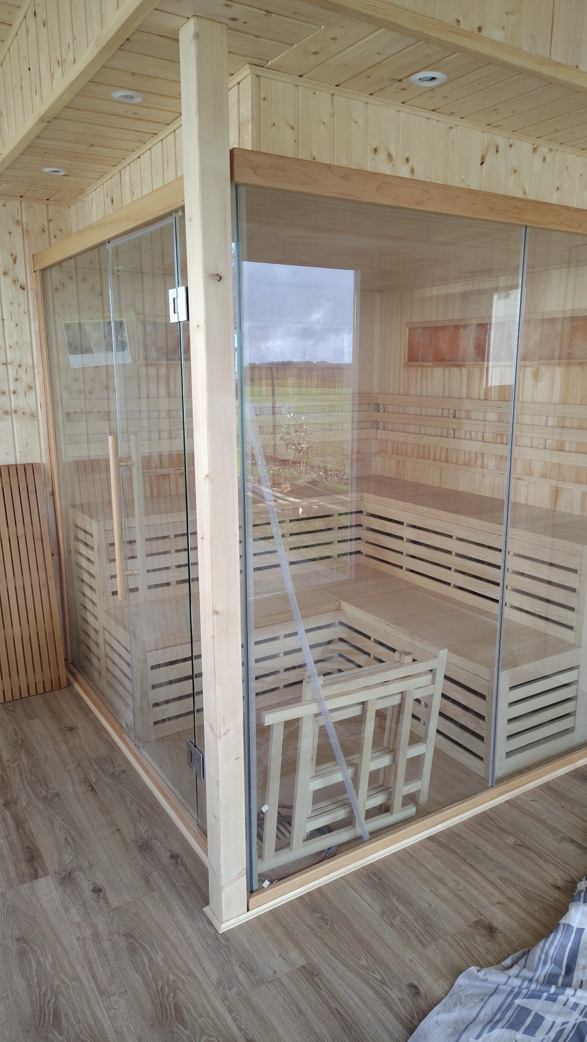 Nowoczesna sauna z jasnego drewna i przeszklonymi ścianami, widok z zewnątrz, ukazujący wnętrze z ławkami i oparciami. Widoczne elementy konstrukcyjne i wykończeniowe.