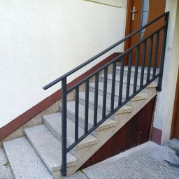 Tomasz Sekuła MetalFusion - Zewnętrzne schody z szarego lastryko, zabezpieczone prostą, ciemną metalową poręczą. Widoczny fragment elewacji budynku i drzwi wejściowe.