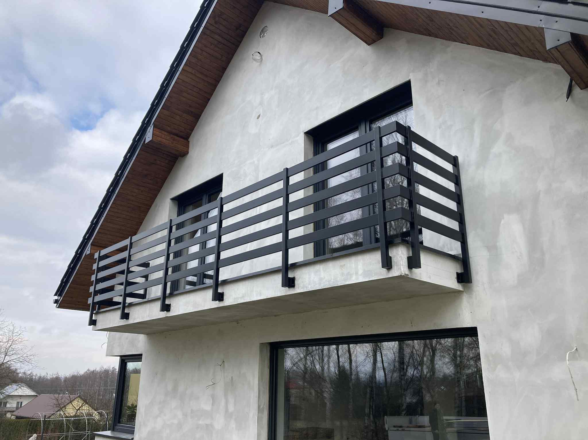 Czarna, pozioma balustrada balkonowa z metalu, zamontowana na balkonie domu z szarą elewacją. Widok z dołu, podkreślający nowoczesny design i solidne wykonanie.