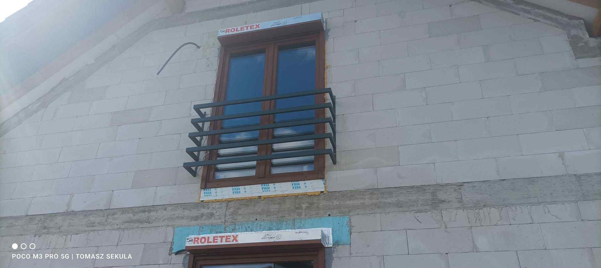 Surowy stan budynku z oknami i metalową balustradą balkonową. Widoczne bloczki betonowe i elementy montażowe okien. Balustrada w kolorze ciemnoszarym.