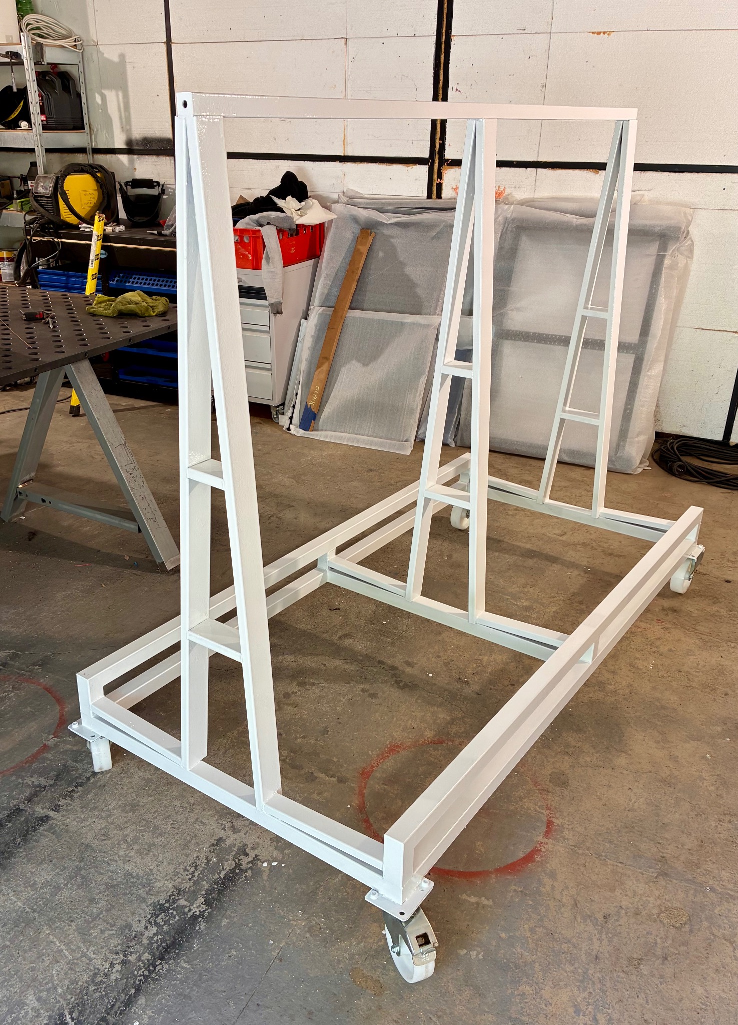 Biała, metalowa konstrukcja na kółkach, prawdopodobnie w trakcie produkcji, widoczna w warsztacie. Elementy spawane, pomalowane na biało. W tle narzędzia i materiały.