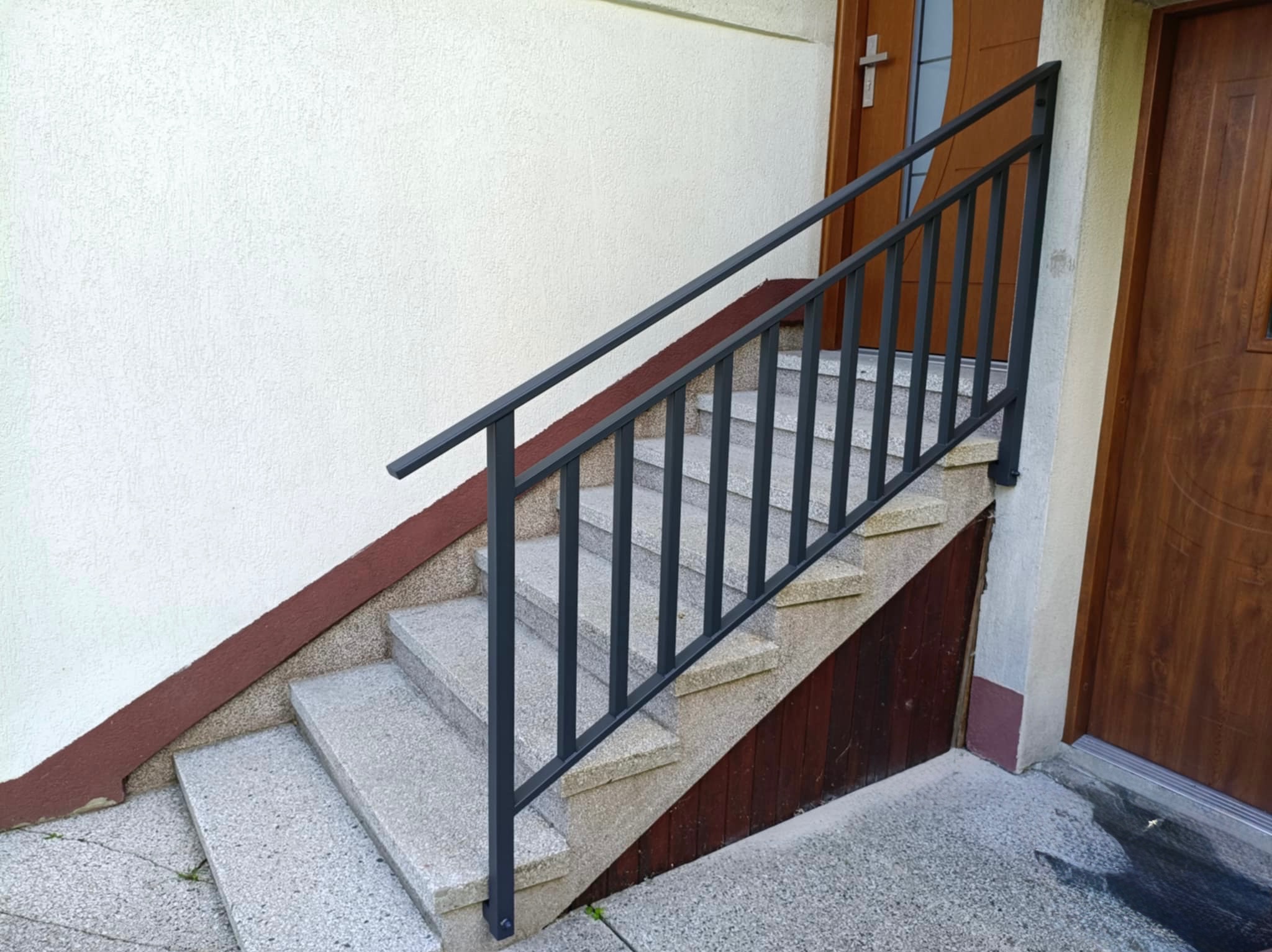 Zewnętrzne schody z szarego lastryko, zabezpieczone prostą, ciemną metalową poręczą. Widoczny fragment elewacji budynku i drzwi wejściowe.