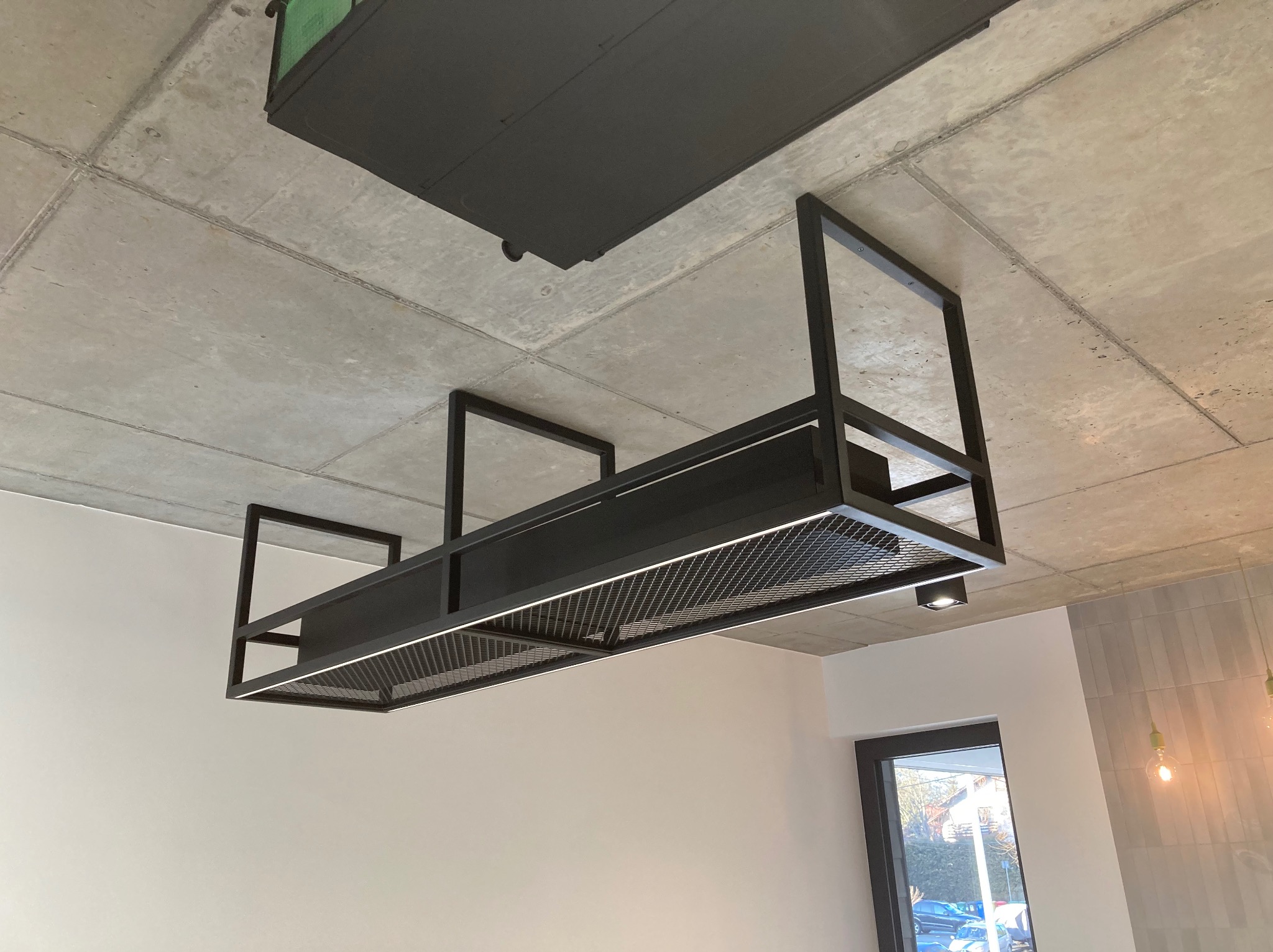 Nowoczesna, czarna, metalowa konstrukcja sufitowa z oświetleniem LED, widoczna na tle surowego, betonowego sufitu i białych ścian. Minimalistyczny design.