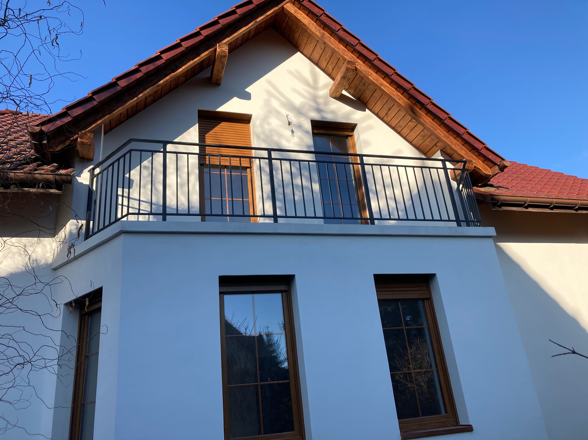 Biały dom z balkonem i czarną, metalową balustradą. Widoczne okna z drewnianymi ramami i czerwony dach. Ujęcie z dołu, w słoneczny dzień.
