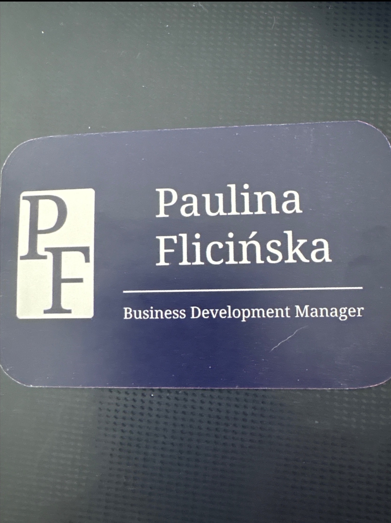 Wizytówka Pauliny Flicińskiej, Business Development Manager, z logo PF na granatowym tle, leżąca na ciemnej powierzchni z delikatną fakturą.