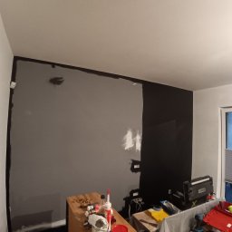 ANDRZEJ DAMPC SZYBKA USTERKA I REMONT - Ściana w trakcie malowania na szaro i czarno, widoczne narzędzia malarskie, częściowo pomalowana ściana, listwy przypodłogowe, okno balkonowe.